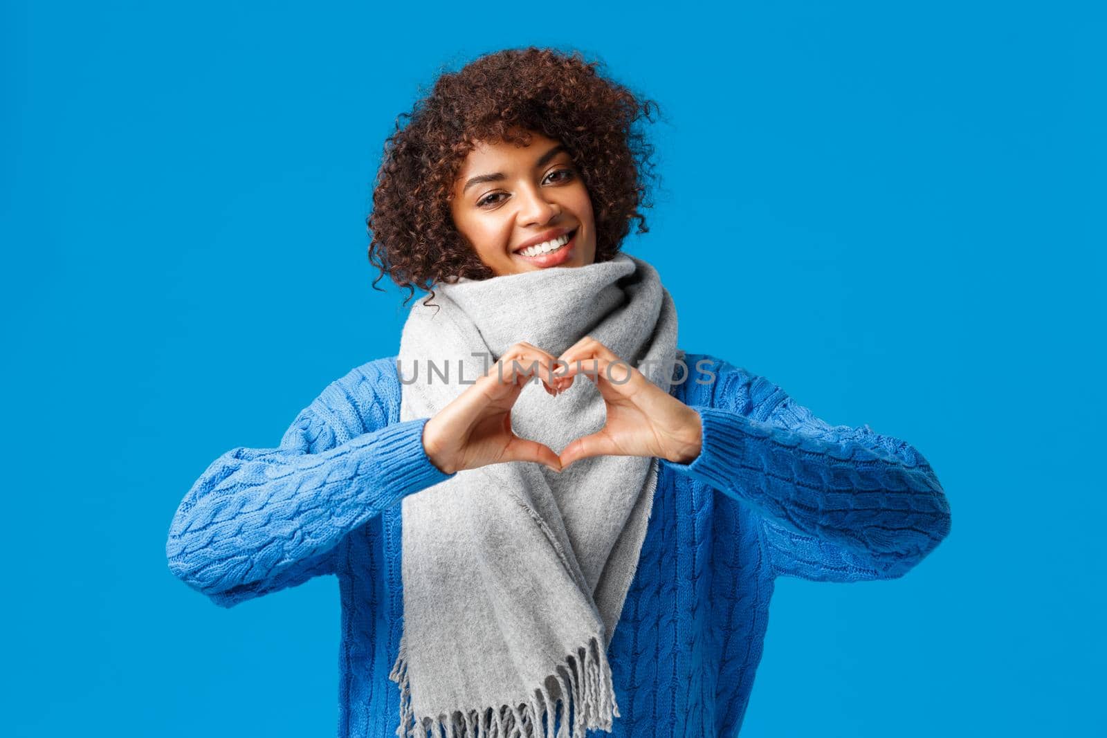 Lovely, romantic cheerful african-american girlfriend with afro haircut, tilt head showing heart sign, confessing love and affection, wearing winter scarf, sweater, standing over blue background.