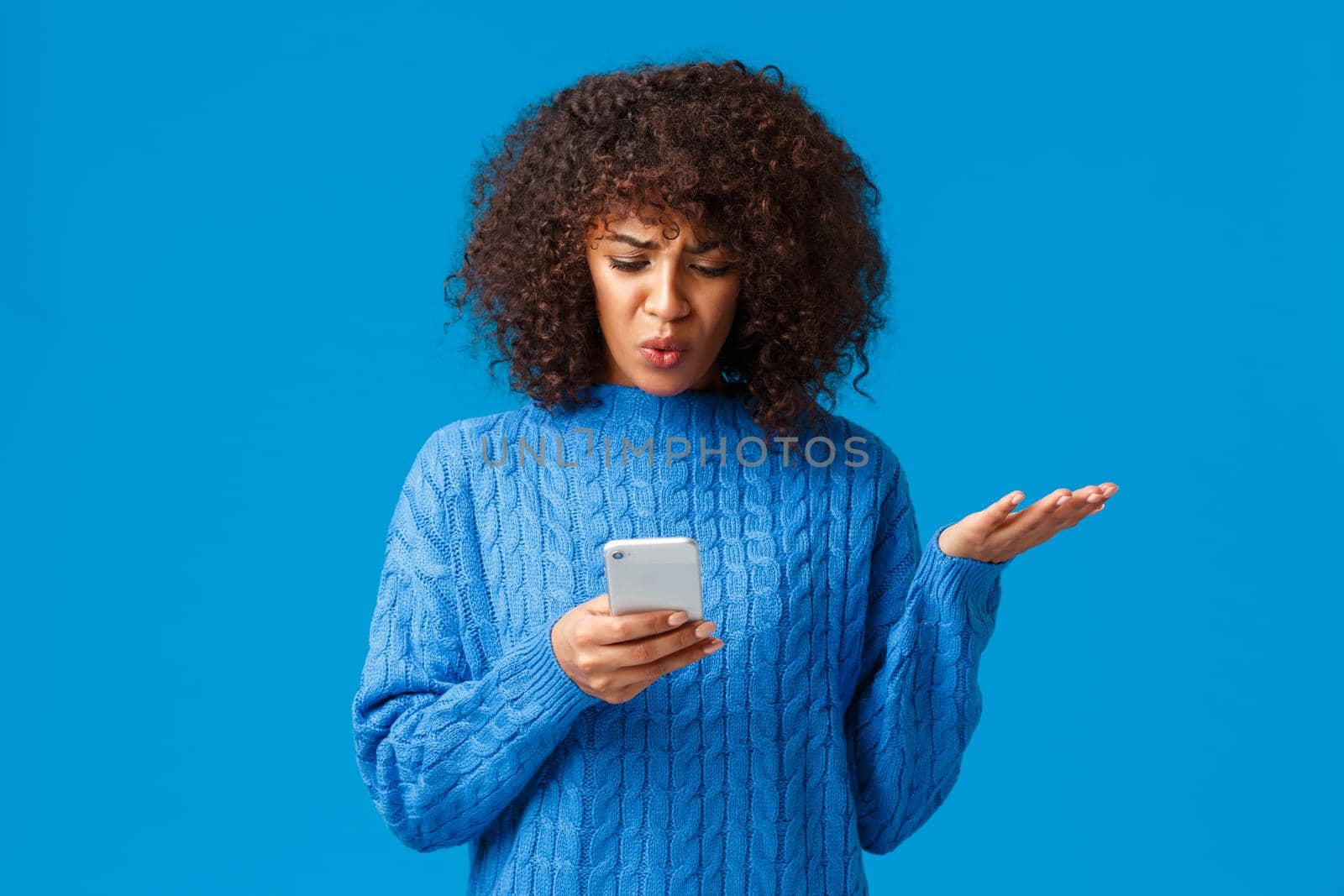 Problem, messaging and people concept. Confused and displeased, tensed african-american young woman receive strange message, staring at smartphone screen clueless, shrugging, blue background by Benzoix