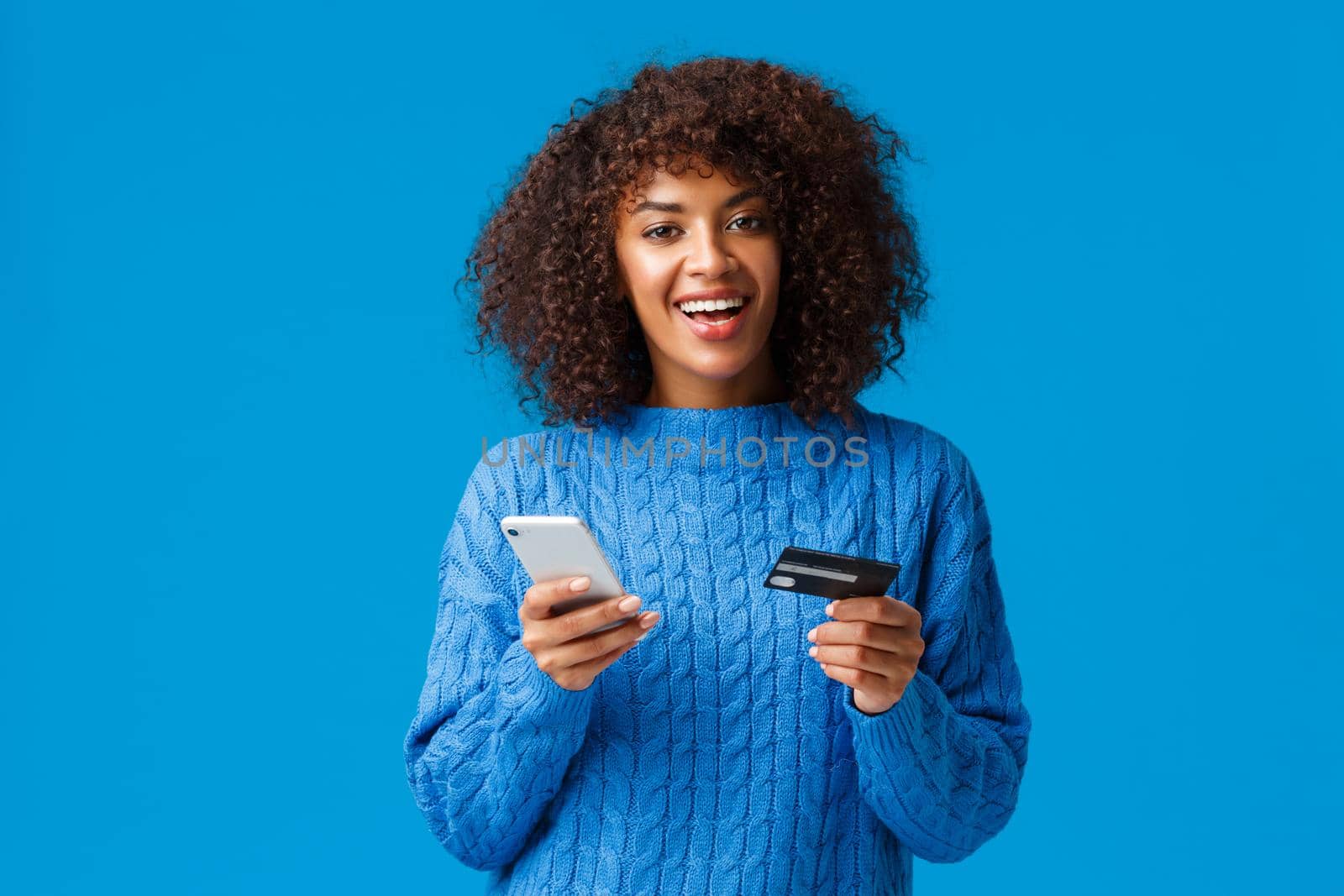 Cheerful good-looking smiling african-american woman buying online, shopping during discounts holiday season, smiling, holding smartphone and credit card, standing blue background.