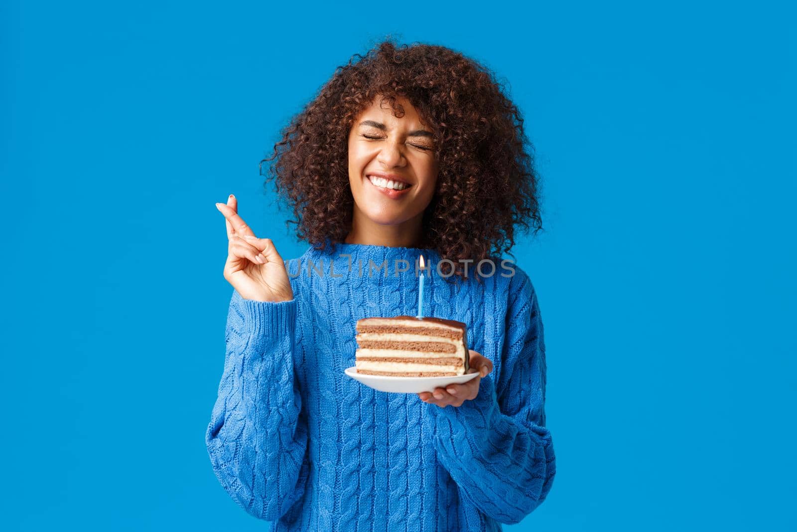 Hopeful and excited, cute african-american woman in sweater, making wish and cross fingers good luck, hope dream come true as close eyes and blowing-out candle on birthday cake.