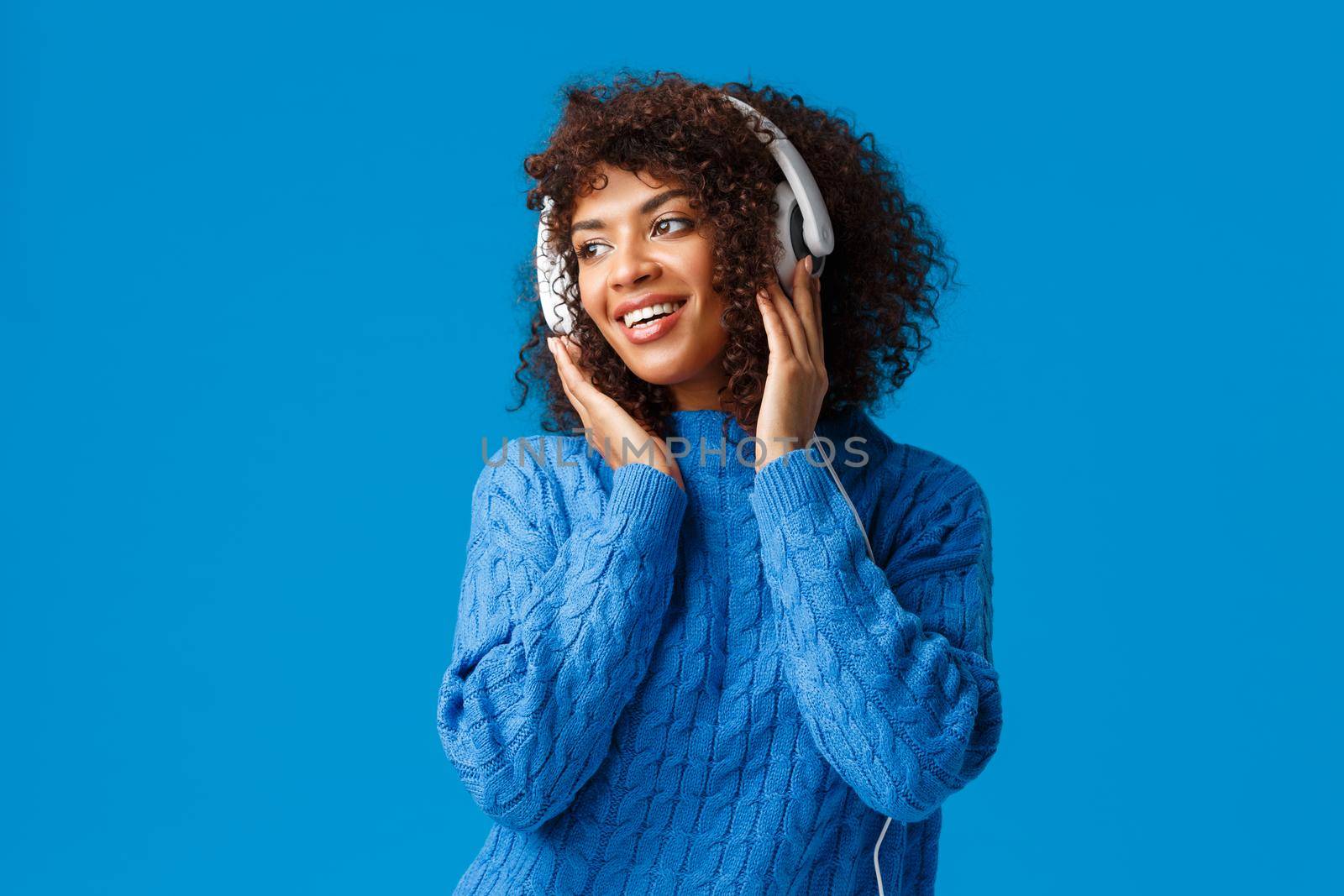 What lovely romantic music. Attractive sensual african-american feminine girl with afro haircut, wearing winter sweater, looking left with pleasant smile, wearing headphones, listen songs by Benzoix