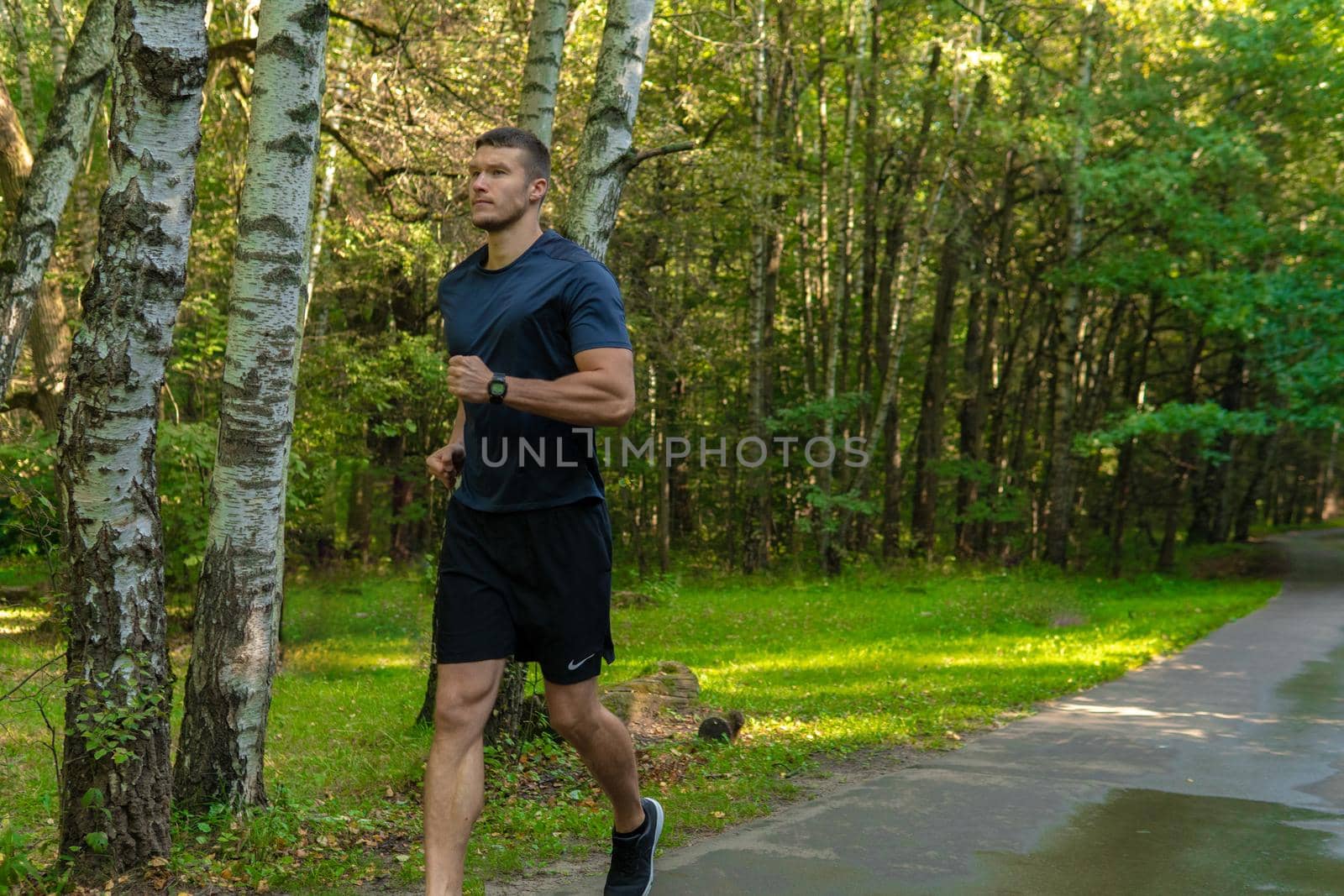 A man athlete runs in the park outdoors, around the forest, oak trees green grass young enduring athletic athlete runner exercise, workout training person jog, trees outside. Summer energy cross, feet stretches