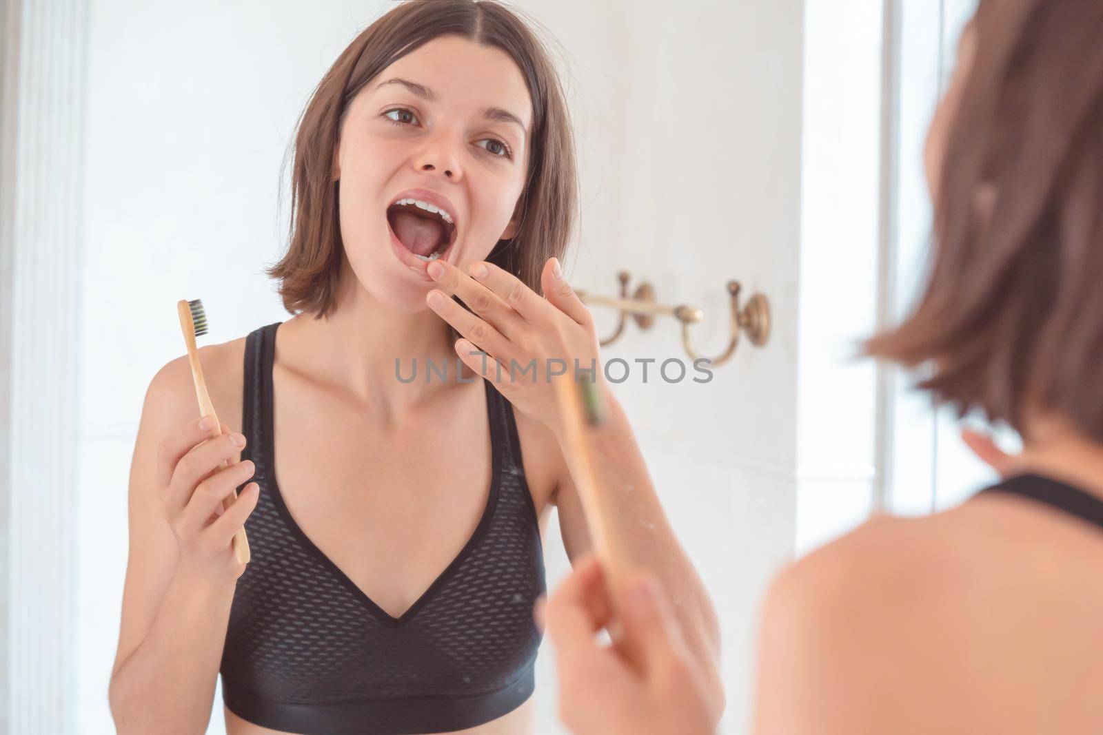 A young smiling girl brushes her teeth with toothpaste in the bathroom by the mirror in the morning, a woman takes good care of the health of tooth enamel and gums.