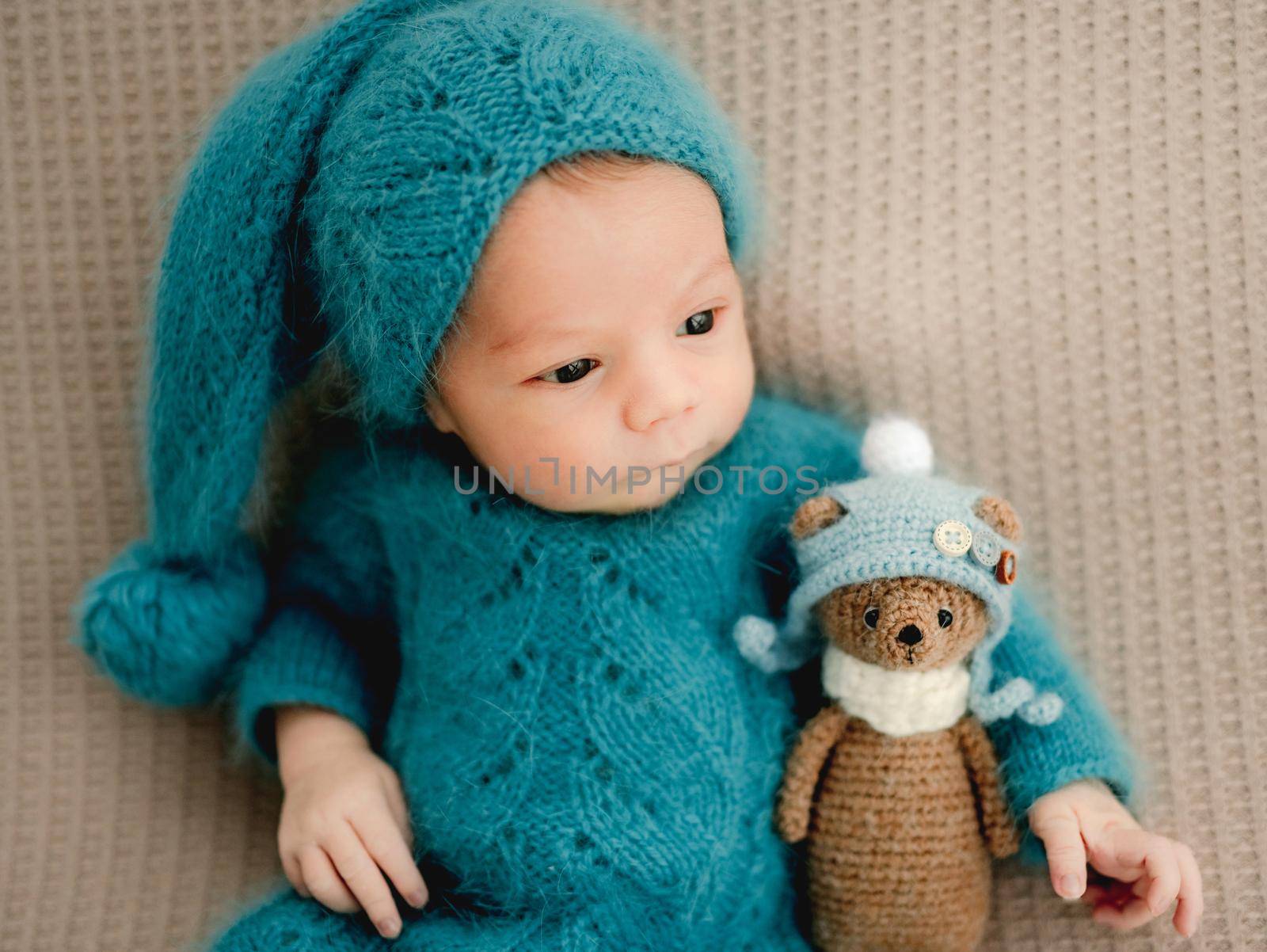 Newborn baby boy wearing knitted costume and hat holding teddy bear toy and looking back. Cute infant child kid studio portrait