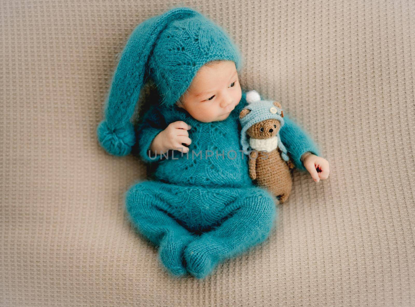 Newborn baby boy wearing knitted costume and hat holding teddy bear toy and looking back. Cute infant child kid studio portrait
