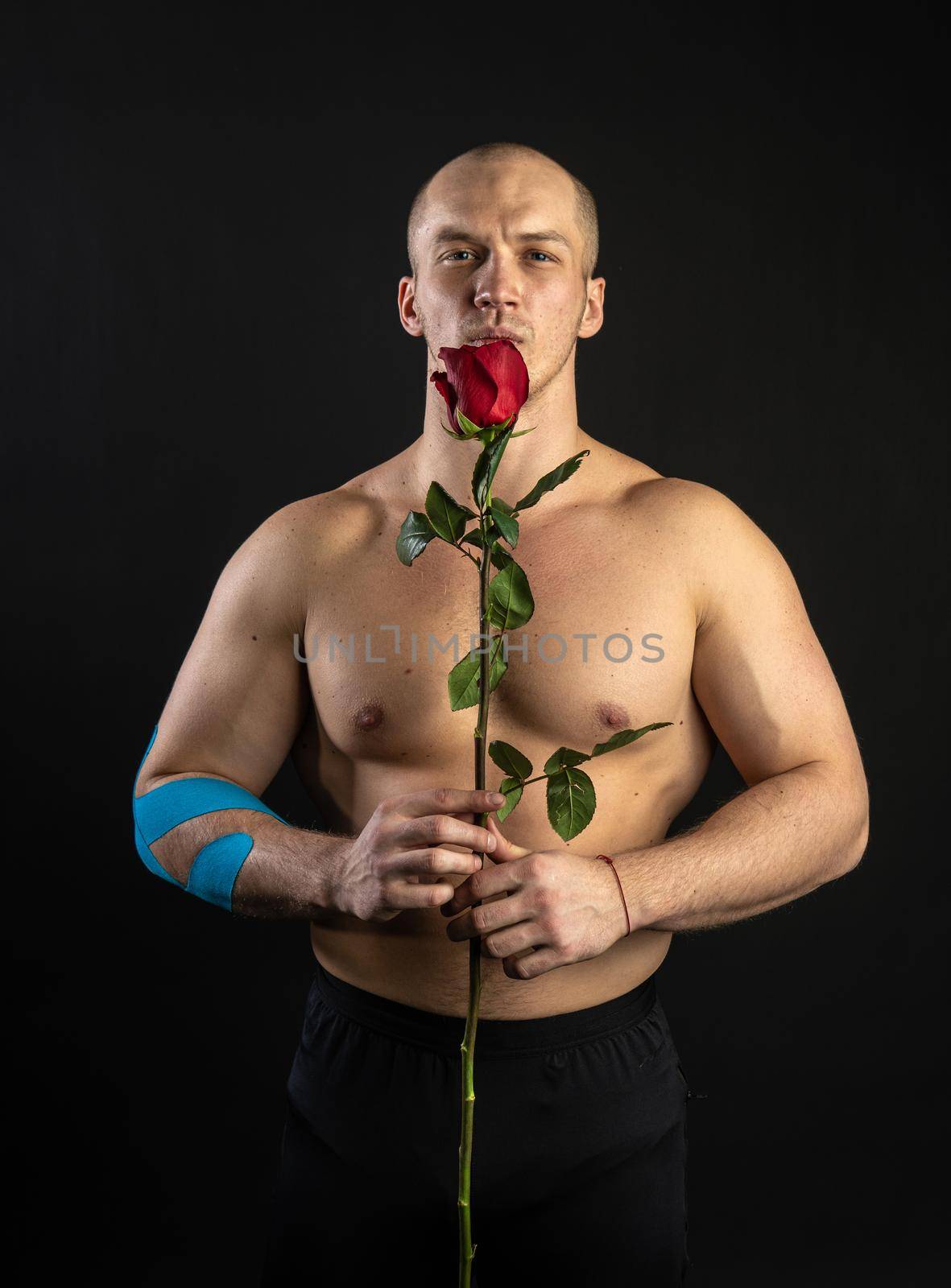 Athletic man holding a red rose, naked for the day of velentin pumped up, on his arm a teip of a bodybuilder romantic. lover guy LGBT. In the studio on a black background Joyful looks at the camera
