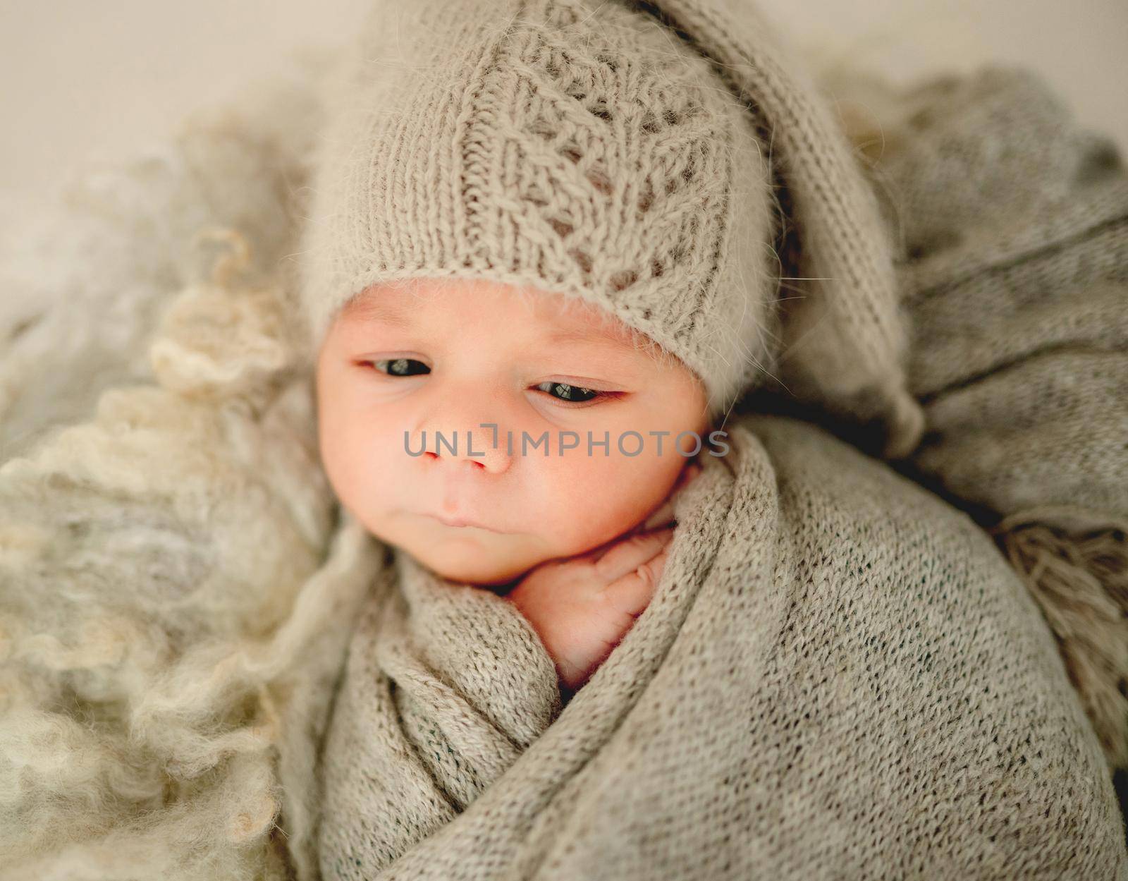 Newborn baby boyvswaddled in knitted fabricand wearing hat falling asleep. Cute infant child kid closeup studio portrait