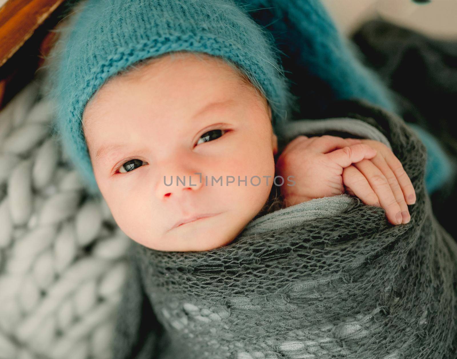 Newborn baby boy swaddled in fabric and wearing knitted hat looking at camera. Cute infant child kid studio portrait