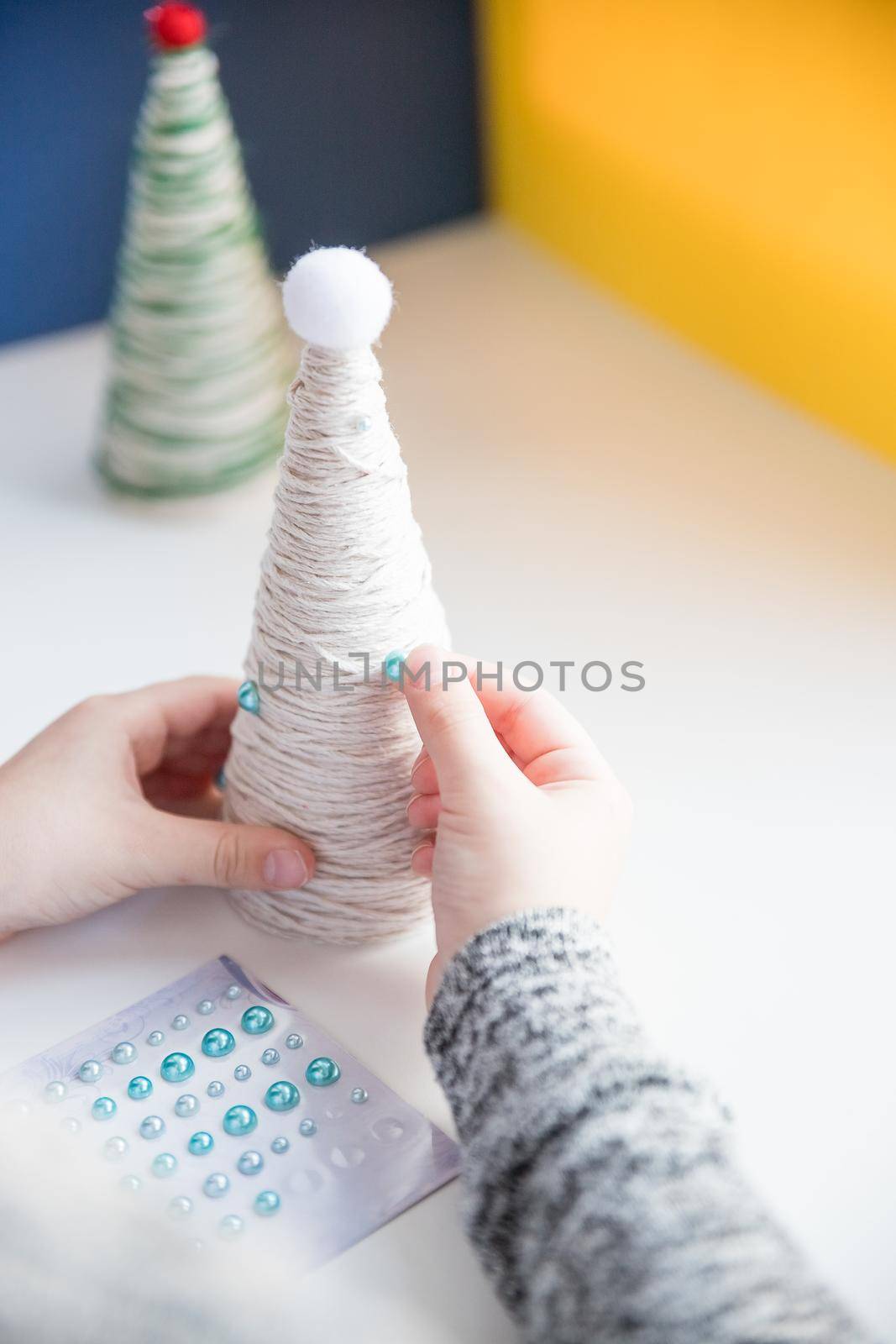 Kid is holding a Christmas tree decoration in his hands. Kid is showing a Christmas tree decoration. Merry Christmas tree project for kids. Scissors, green cotton yarn on a wooden table