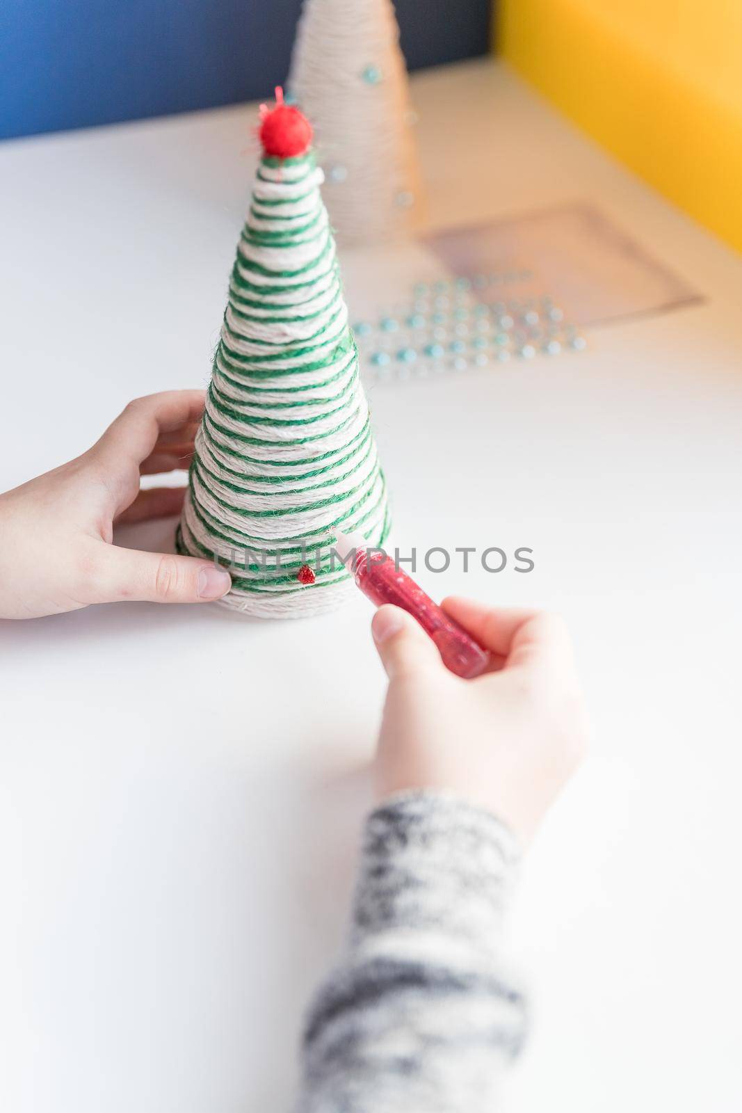 Kid is holding a Christmas tree decoration in his hands. Kid is showing a Christmas tree decoration. Merry Christmas tree project for kids. Scissors, green cotton yarn on a wooden table