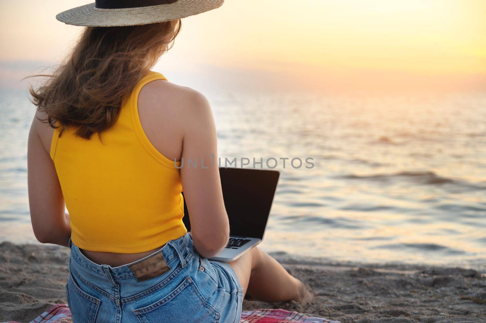 View from the back. Caucasian woman uses and types on the keyboard of a laptop computer while sitting on a beautiful beach at sunset.