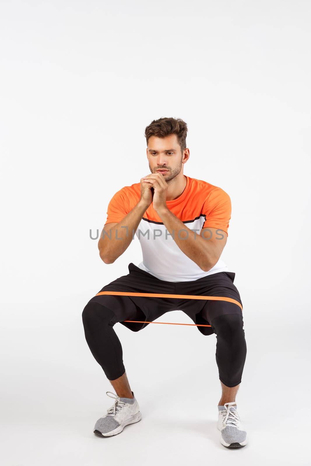 Motivated, focused good-looking sportsman perform squats with resistance rope, stretching equipment by bending feet sideways, sit and clench arms together to be good shape, workout concept.
