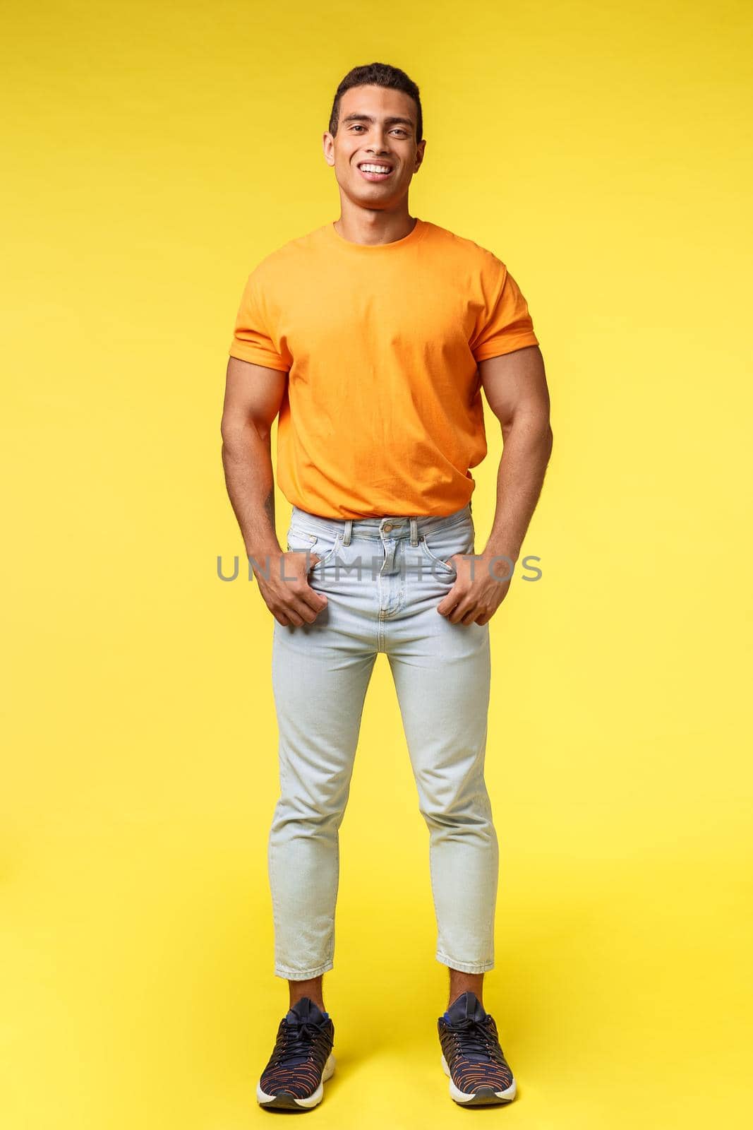 Vertical full-length studio shot young hipster guy with mascular body, standing orange t-shirt and white pants, hold hands in pockets, smiling camera pleased and confident, casual pose.