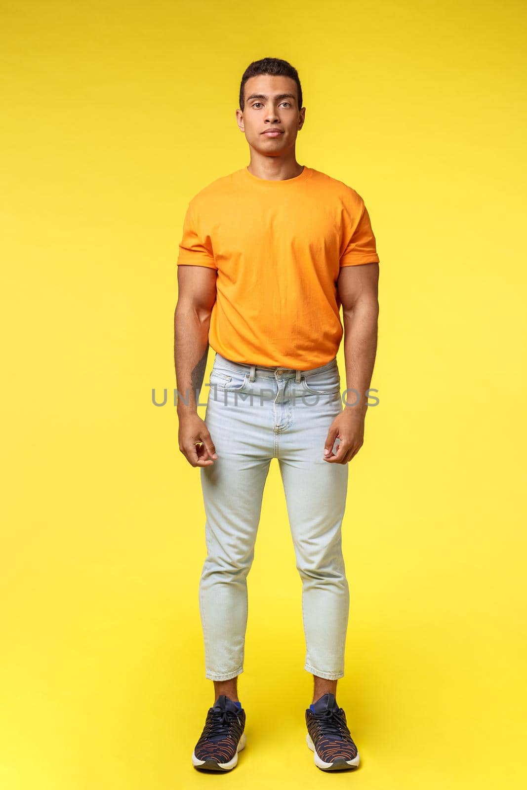 Full-length vertical shot masculine hipster guy, hispanic ethnicity, standing casually yellow background, wear stylish orange t-shirt, white pants, look camera with no expression, slightly smiling.