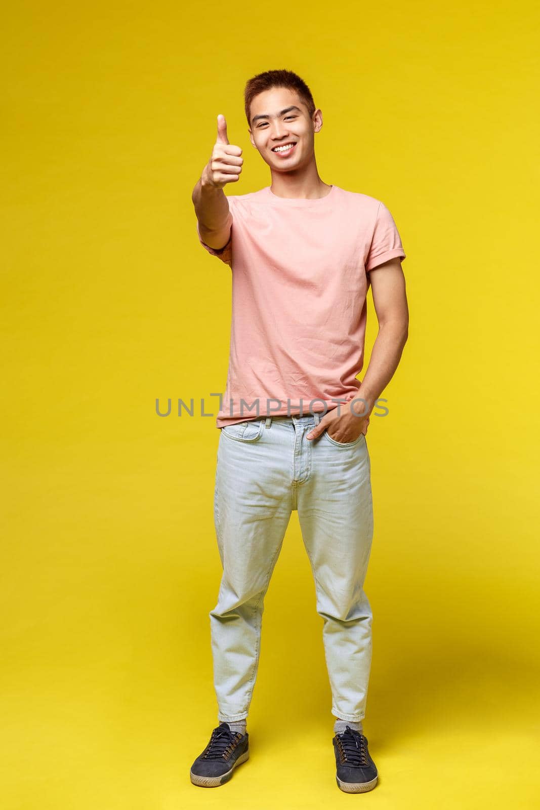 Lifestyle, education and people concept. Vertical portrait of stylish asian hipster guy, show thumb-up and smiling pleased, recommend product, approve and guarantee great quality, yellow background.