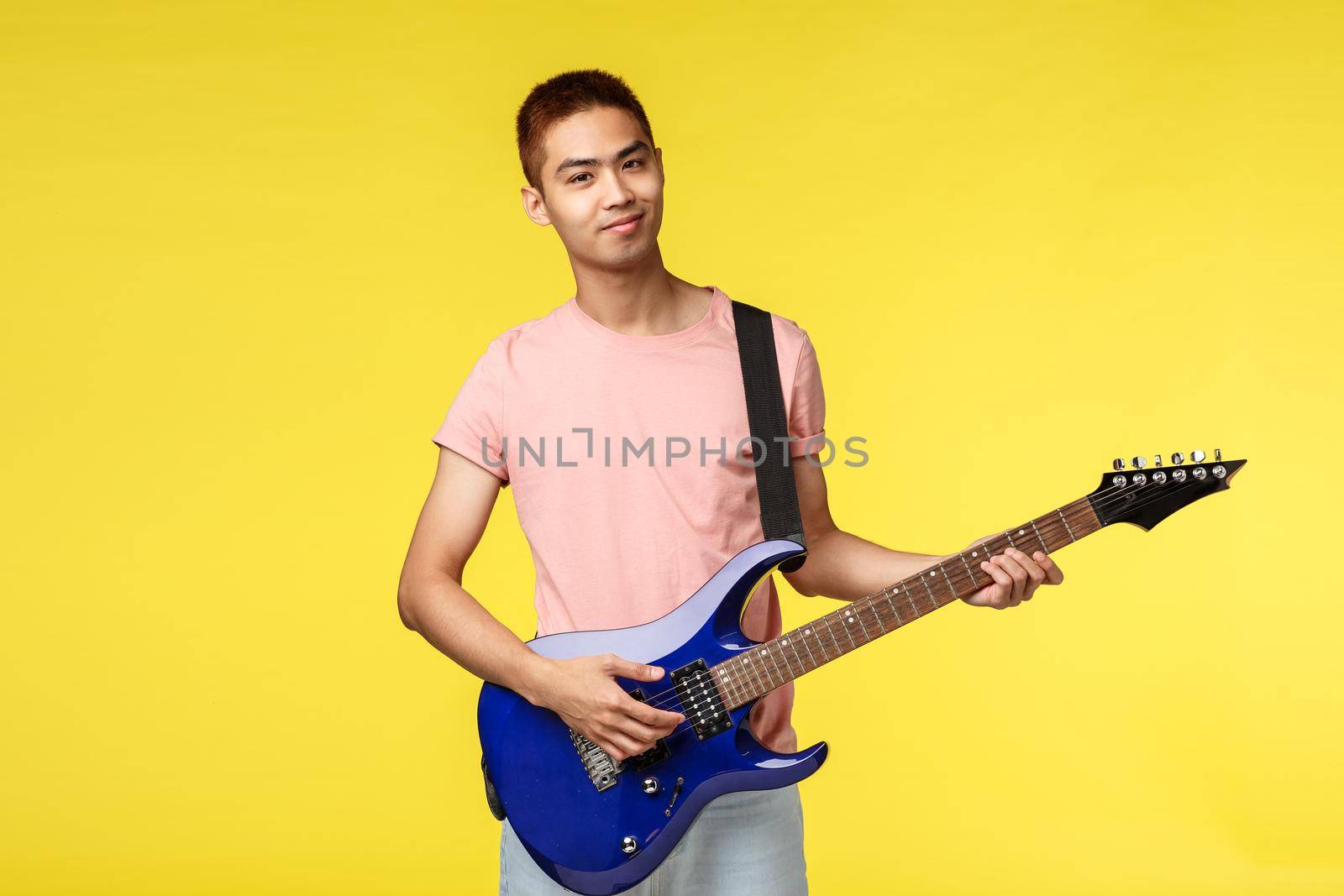 Lifestyle, leisure and youth concept. Portrait of smiling asian young man playing in band, holding electric guitar, practicing music, learning play instrument, stand yellow background.