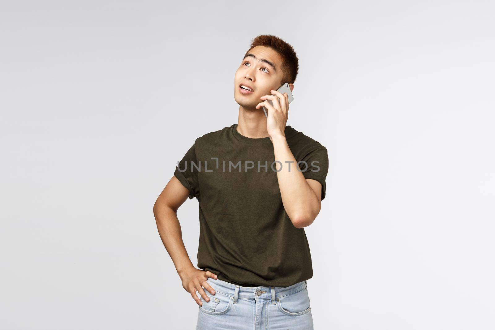 Technology, online lifestyle and communication concept. Thoughtful good-looking asian male talking on mobile phone, look dreamy up, thinking what answer, order food delivery, grey background.