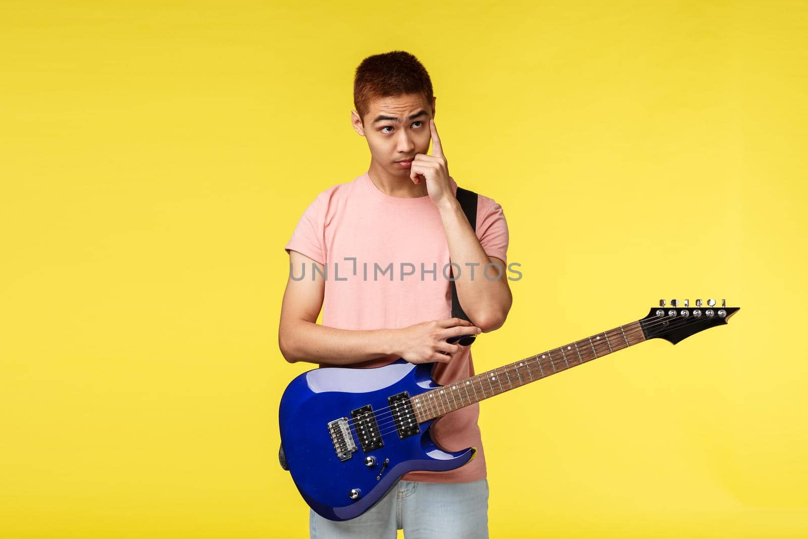 Lifestyle, leisure and youth concept. Thoughtful asian male in pink t-shirt, scratching face and look away with indecisive, puzzled look, holding electric guitar, learn how play, yellow background by Benzoix