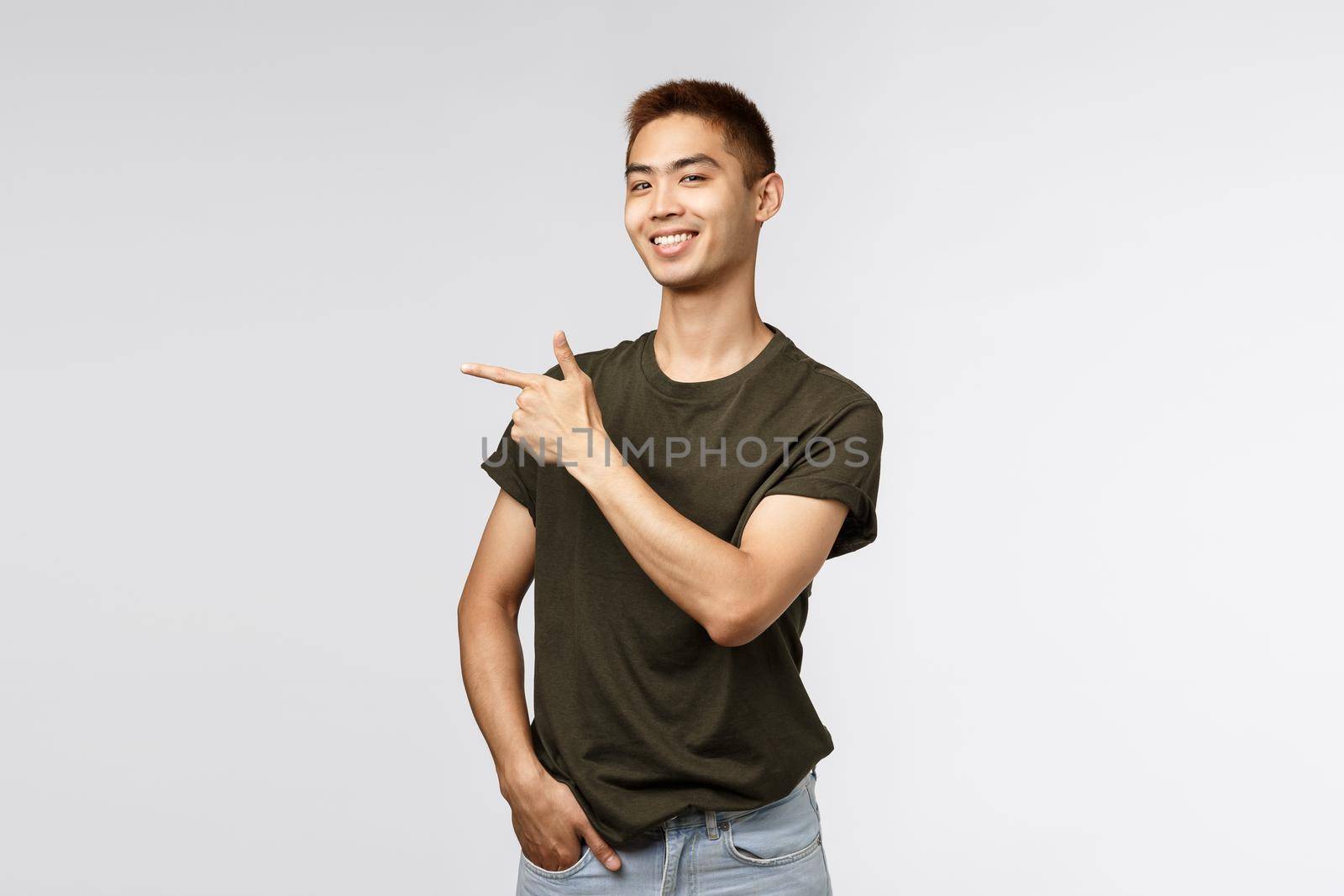 Proud good-looking asian male student pointing finger left and smiling camera pleased, guy showing way to store, online link or company banner, introduce item, grey background.