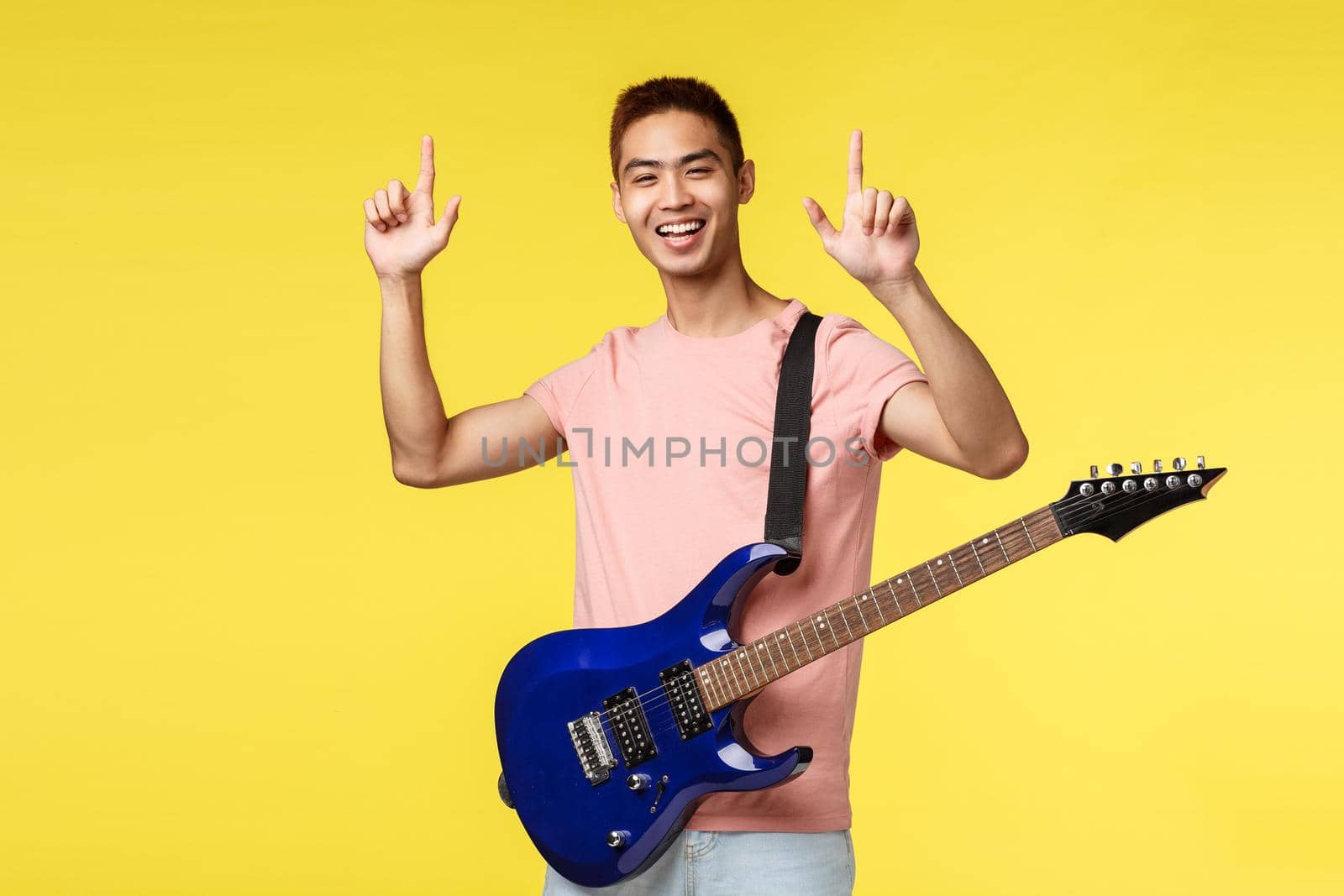 Lifestyle, leisure and youth concept. Happy enthusiastic handsome young asian male pointing fingers up, performing on stage, holding electric guitar and smiling, yellow background.