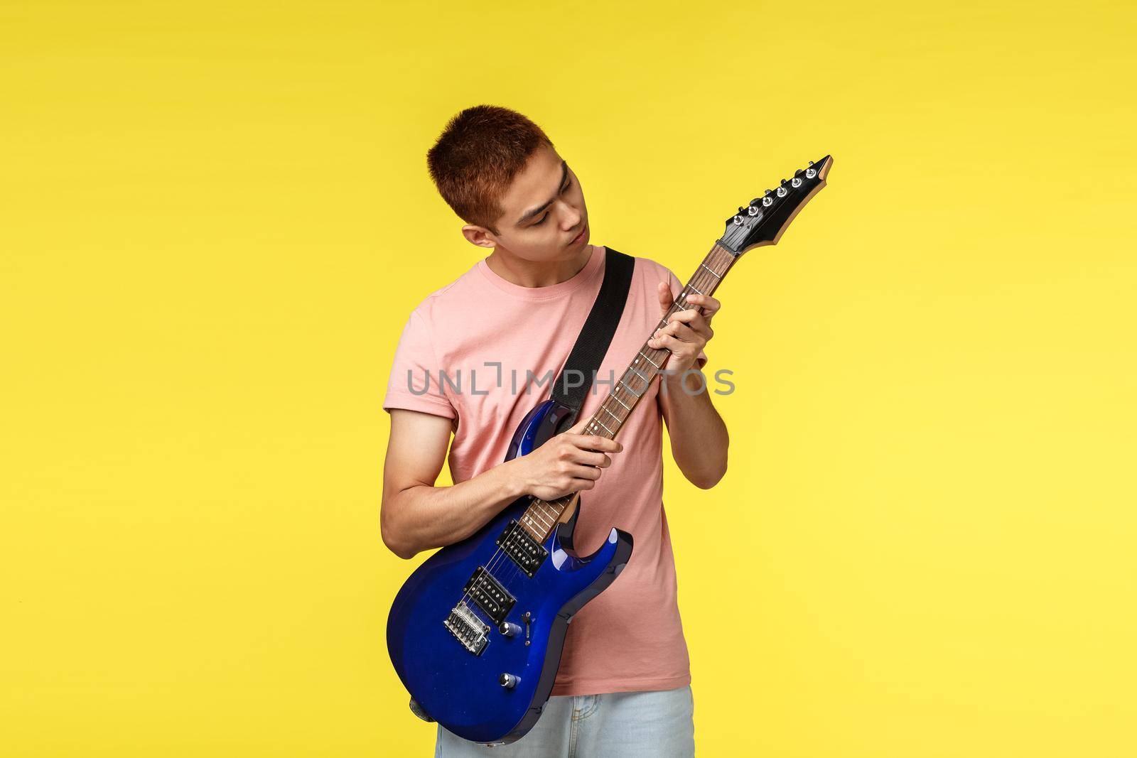 Lifestyle, leisure and youth concept. Serious young handsome man, asian teenage guy playing electric guitar, tune instrument, look at griff curious, stand yellow background, learn new skill.