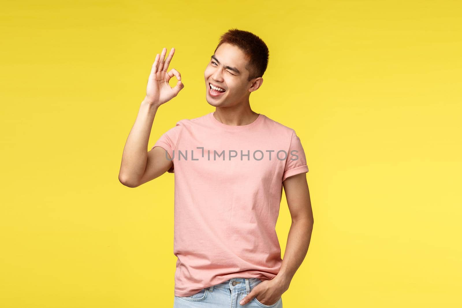 Lifestyle, travel and people concept. Enthusiastic cheerful asian hipster guy guarantee best quality, show okay good sign and wink camera, smiling pleased, recommend item, yellow background.