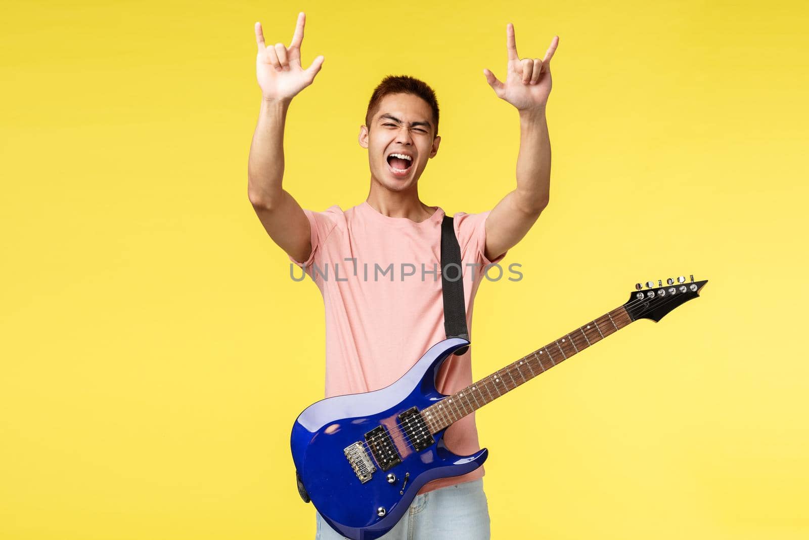 Lifestyle, leisure and youth concept. Good-looking asian guy having fun performing on stage, hold electric guitar, show rock-n-roll gesture and shouting enthusiastic, have fun, yellow background.