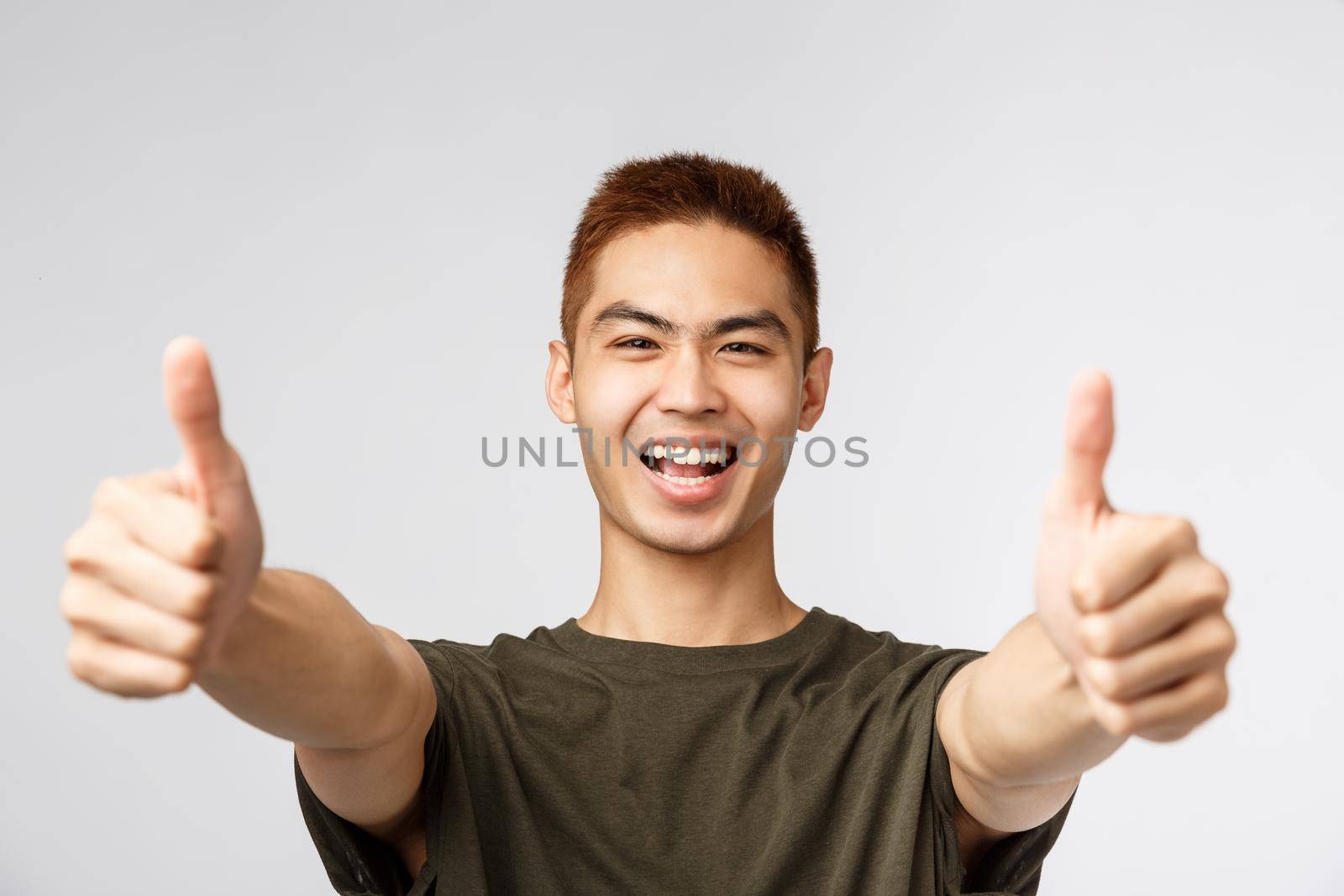 People, different expressions and lifestyle concept. Enthusiastic excited happy man, asian guy show thumbs-up and smiling, saying yes to awesome idea, approve and like concept, grey background by Benzoix