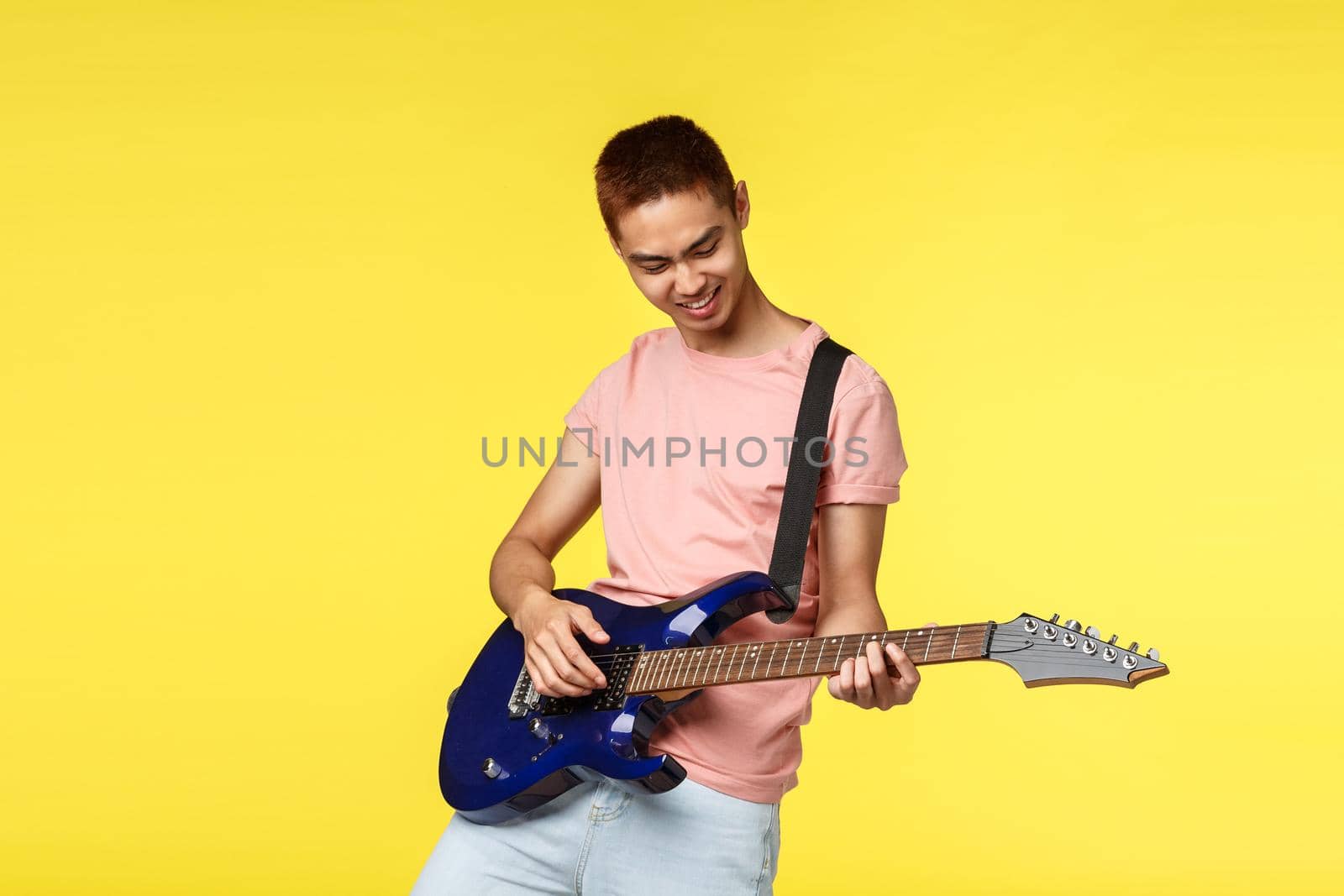 Lifestyle, leisure and youth concept. Portrait of happy, cool and stylish asian guy playing in band, enjoying perfoming on electric guitar, smiling enthusiastic, standing yellow background.