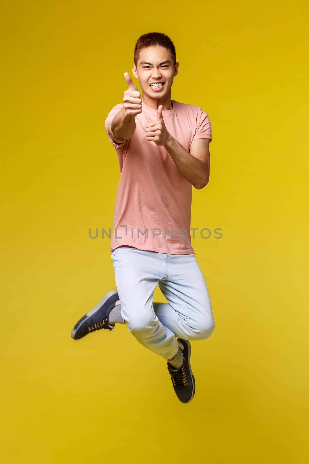 Summer vacation, education and lifestyle concept. Vertical shot of handsome cheeky asian teenage guy vibing, jumping from happiness, pointing at camera and smiling, yellow background.