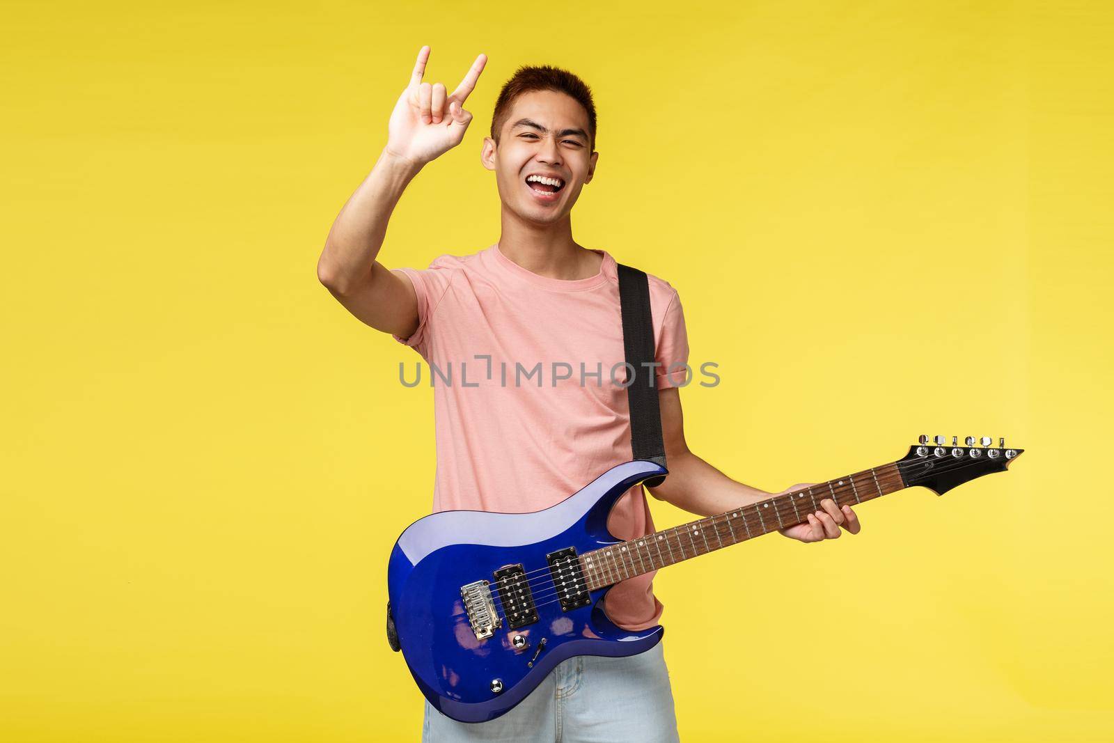 Lifestyle, leisure and youth concept. Happy carefree, good-looking asian guy playing in band, hold electric guitar, show rock-n-roll heavy metal sign and smiling broadly, perform on stage.