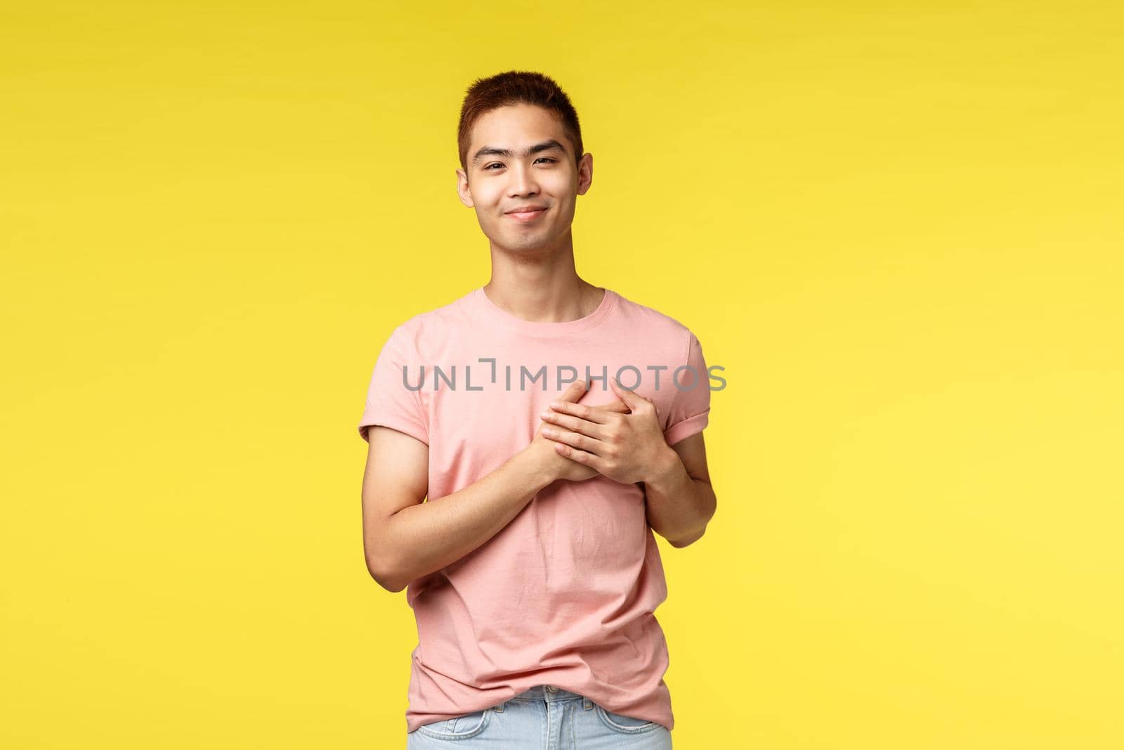 People, different emotions and lifestyle concept. Portrait of touched asian guy holding hands on heart and smiling pleased, keep good memories in soul, being grateful for help, yellow background.