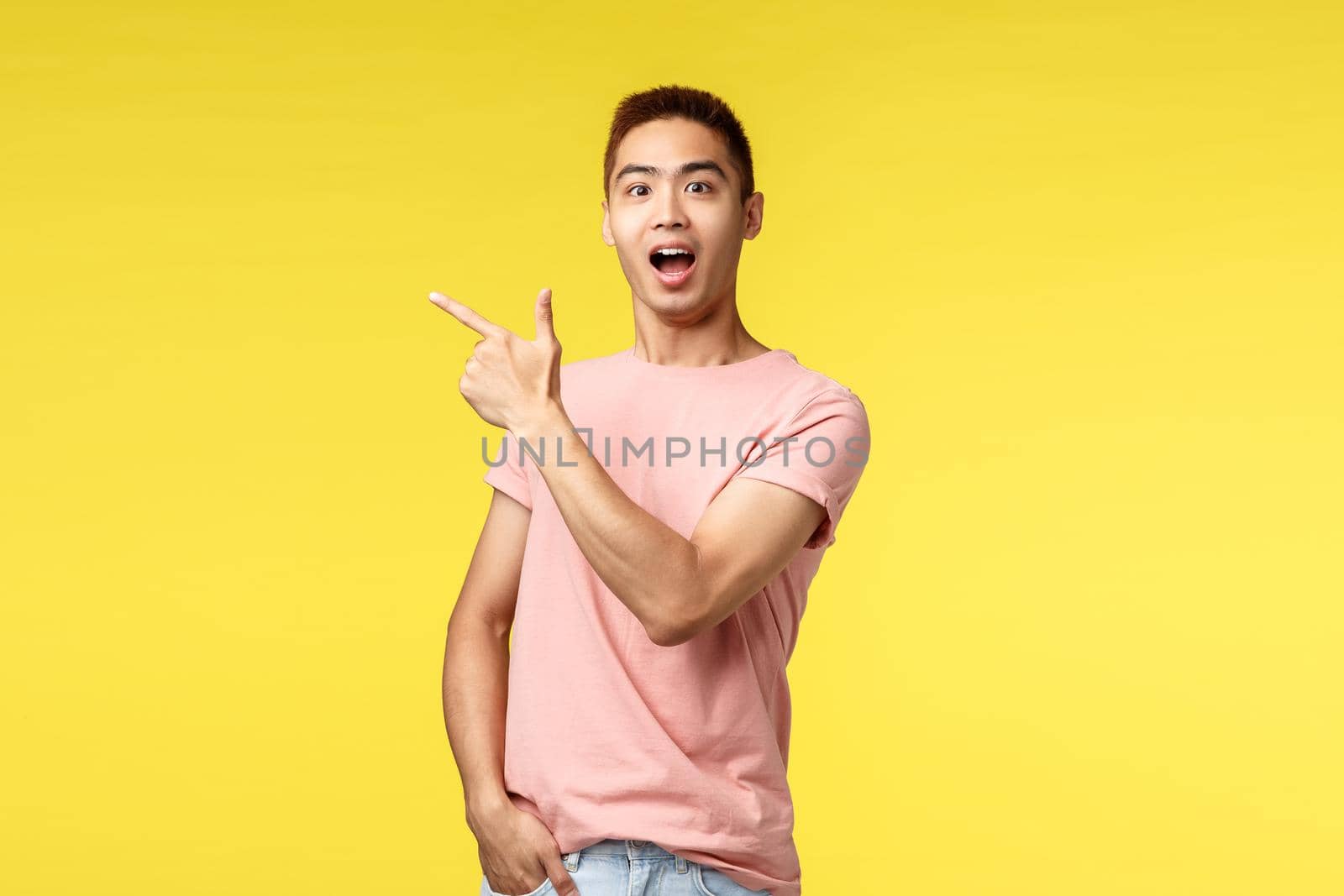 People, different emotions and lifestyle concept. Excited, happy asian man in pink t-shirt showing way, pointing finger upper left corner and smiling amazed, recommend visit store or cafe.