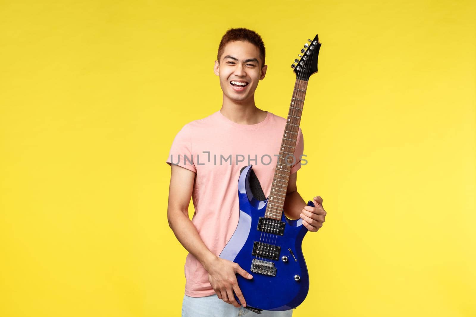 Lifestyle, leisure and youth concept. Enthusiastic handsome guy playing in band, got new electric guitar, laughing and smiling joyful, having fun performing on stage, yellow background.