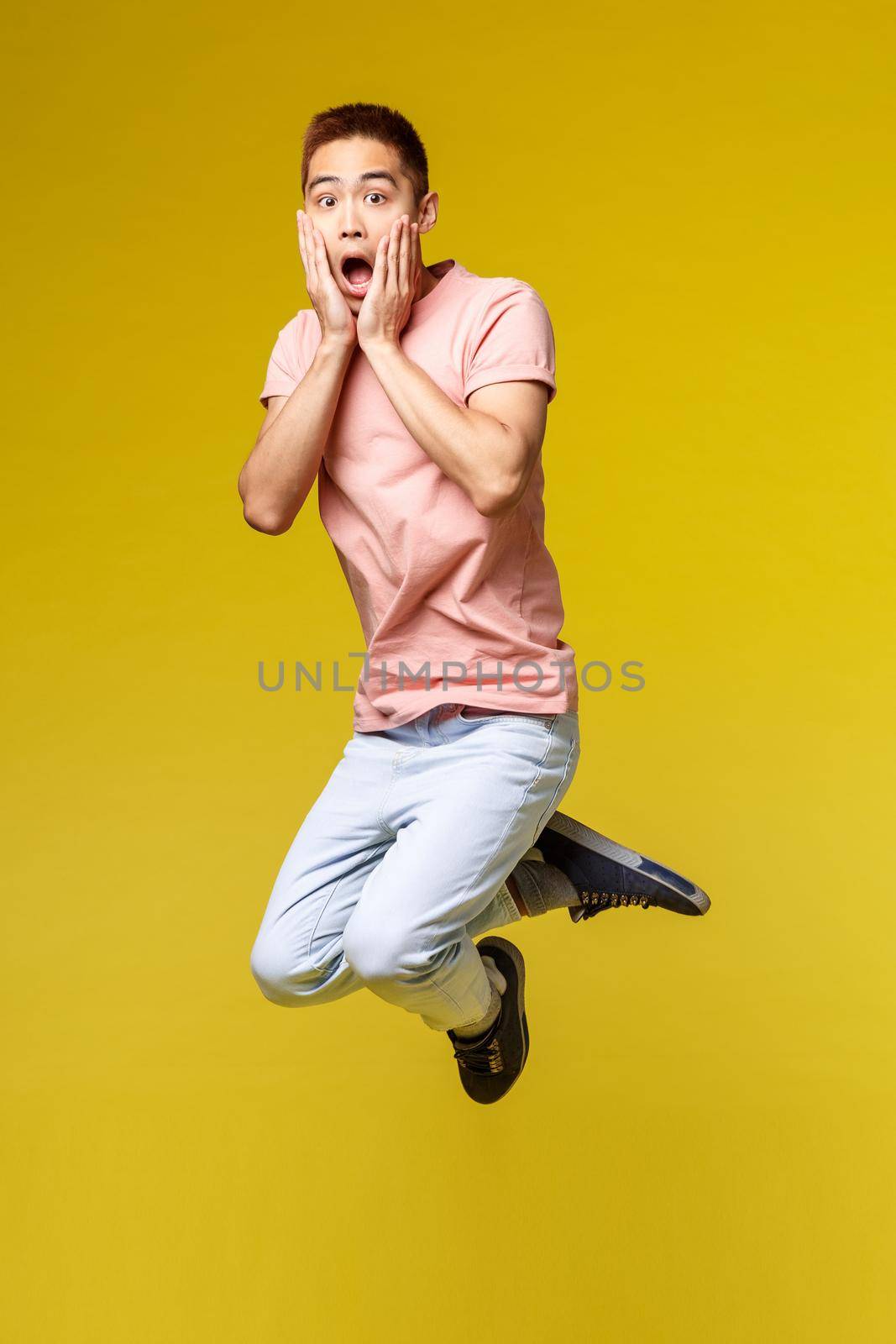 Summer vacation, education and lifestyle concept. Vertical shot of shocked and amazed, gasping asian man heard overwhelming news, touch face and jumping surprised, stand yellow background.