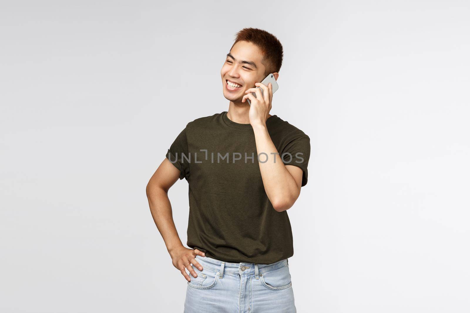 Technology, online lifestyle and communication concept. Handsome cheerful chinese guy in green t-shirt, talking on phone, looking away laughing and smiling upbeat, stand grey background.