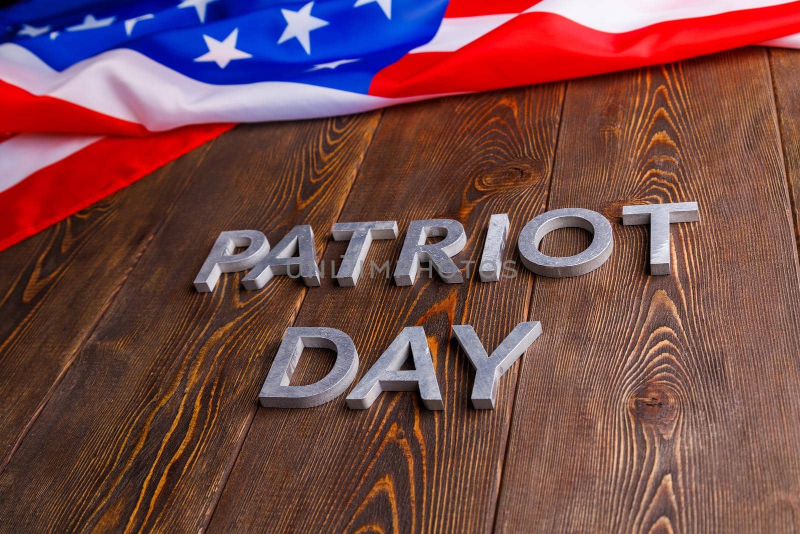 the words patriot day laid with silver metal letters on wooden board surface with crumpled usa flag above in diagonal perspective