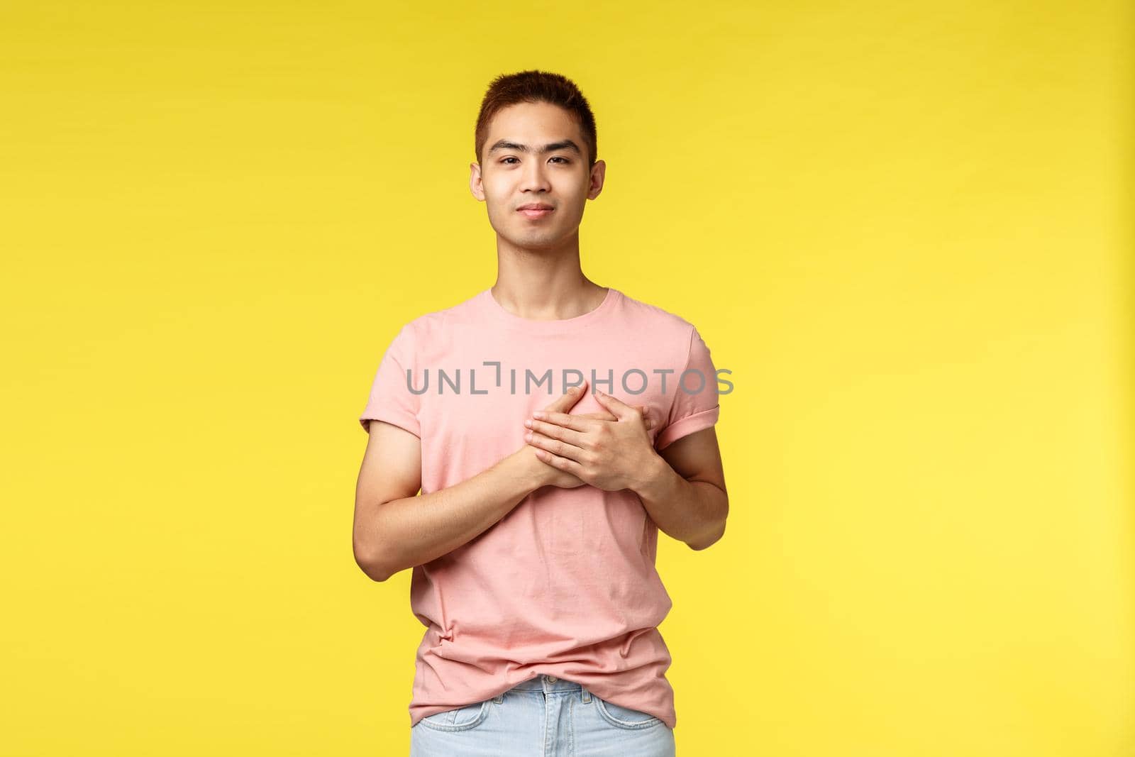 People, different emotions and lifestyle concept. Smiling touched asian man with stylish haircut, pink t-shirt, hold hands on heart and looking camera pleased, going vacation, yellow background.