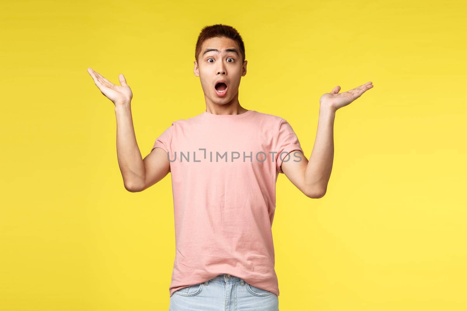 Vacation, summer and people concept. Portrait of surprised handsome, startled asian man gasping, raise hands sideways in amazement and awe, react to wonderful astonishing news, yellow background.