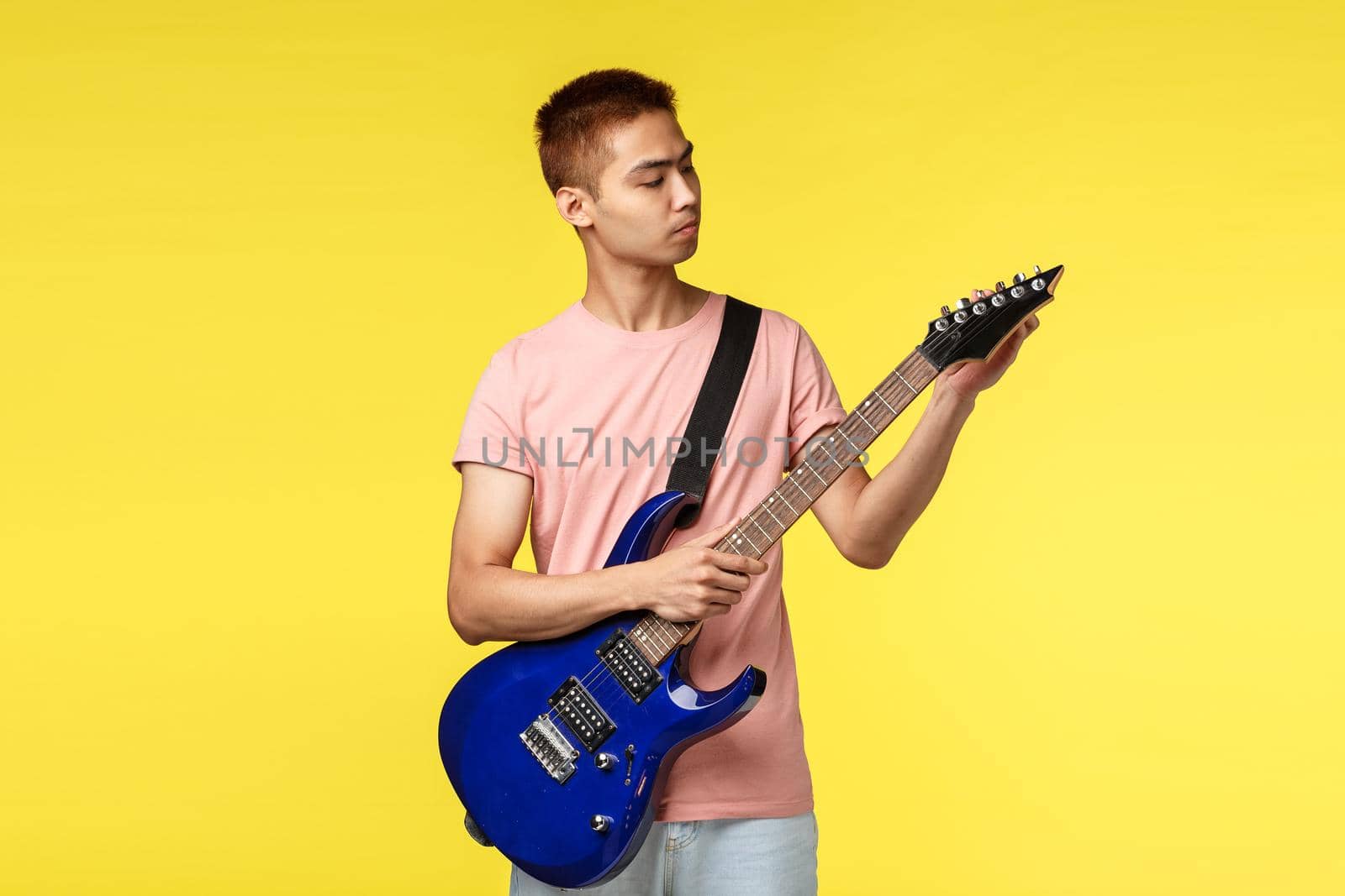 Lifestyle, leisure and youth concept. Portrait of good-looking asian guy playing in band, tune electric guitar before coming on stage and perform song, stand yellow background by Benzoix