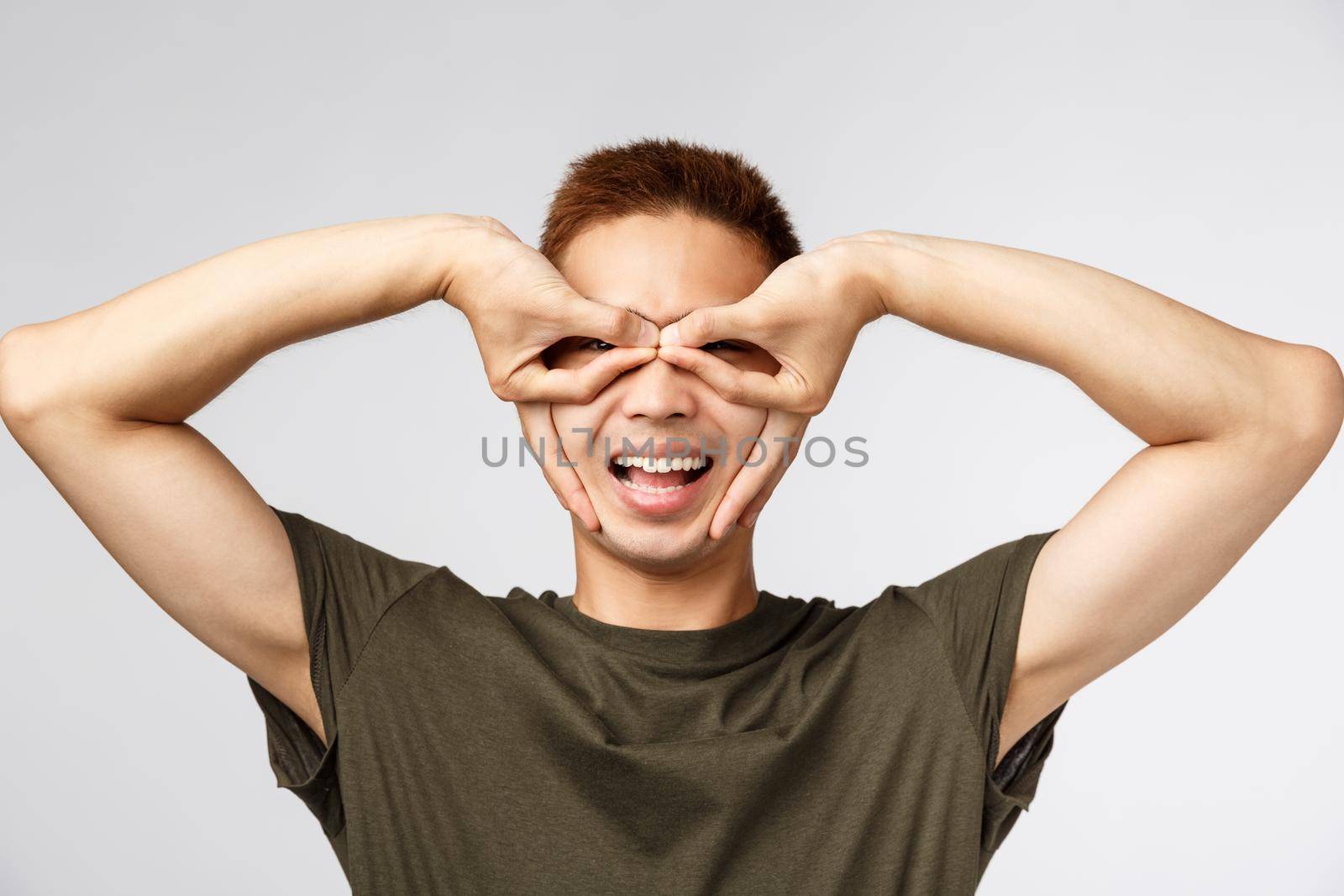 People, different expressions and lifestyle concept. Cheerful good-looking young asian teenage guy having fun, making eye mask with fingers and smiling upbeat, playing around, grey background.