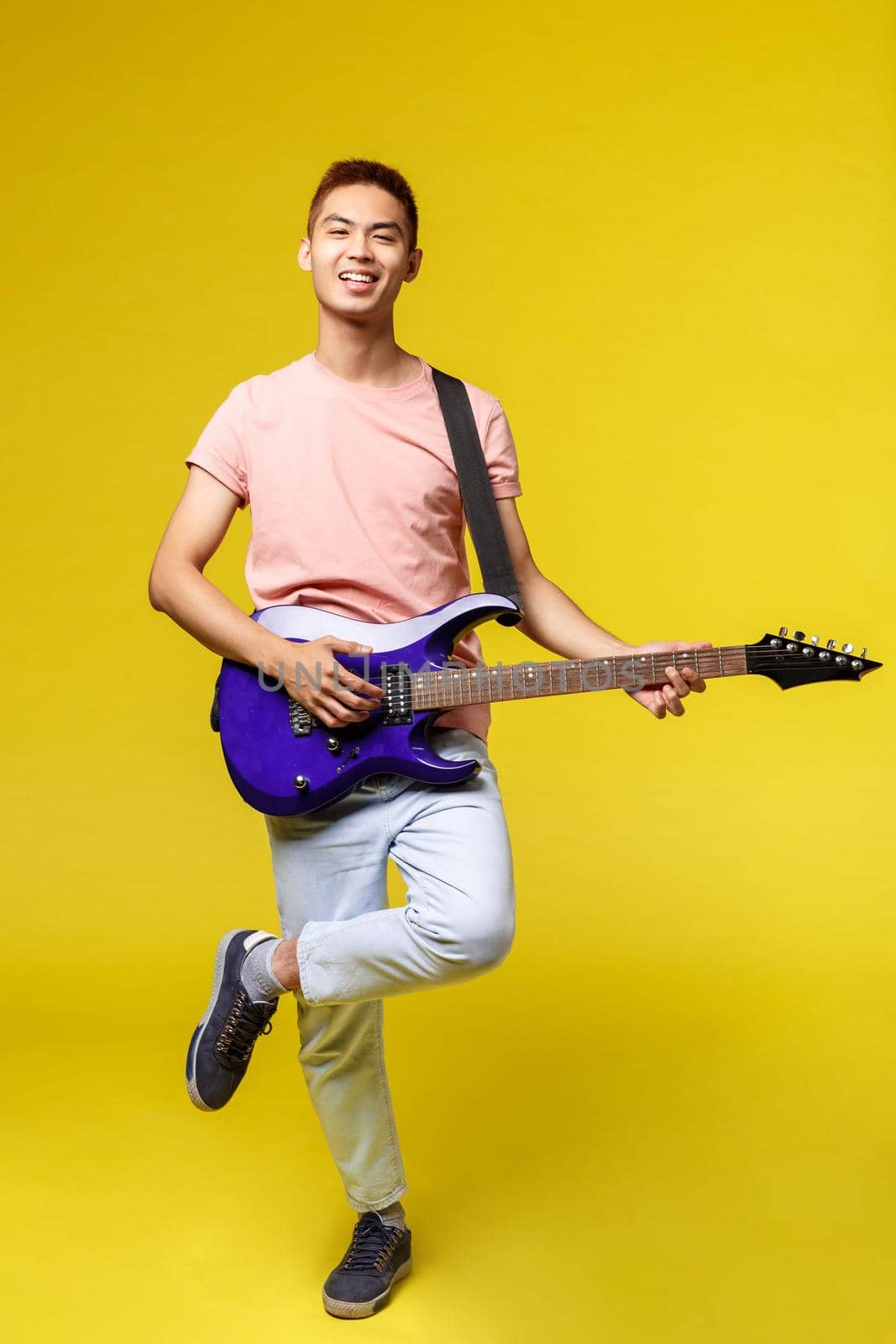 Lifestyle, leisure and youth concept. Vertical portrait of enthusiastic handsome teenage asian guy in pink t-shirt, playing on electric guitar, smiling happy, standing yellow background.