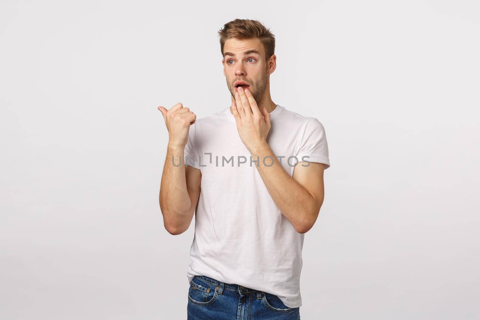 Surprised and shocked young blond handsome guy, ponting thumb left and staring, gasping cover mouth as gossiping, witness something astounding, shocking scene, standing white background.