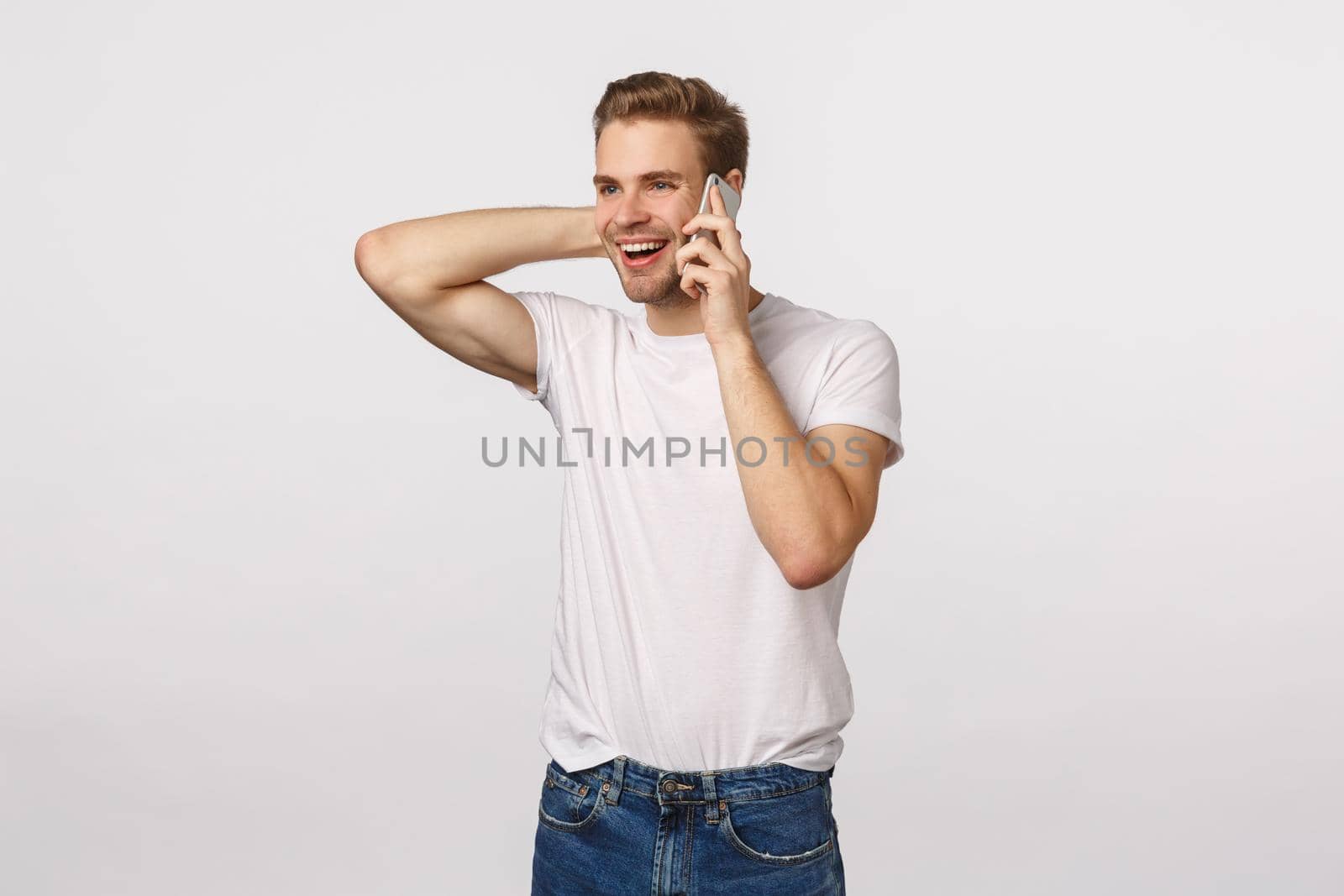 Happy flattered and pleased handsome blond guy receive praises or good news via phone call, holding smartphone near ear, touching neck silly and embarassed, standing white background.
