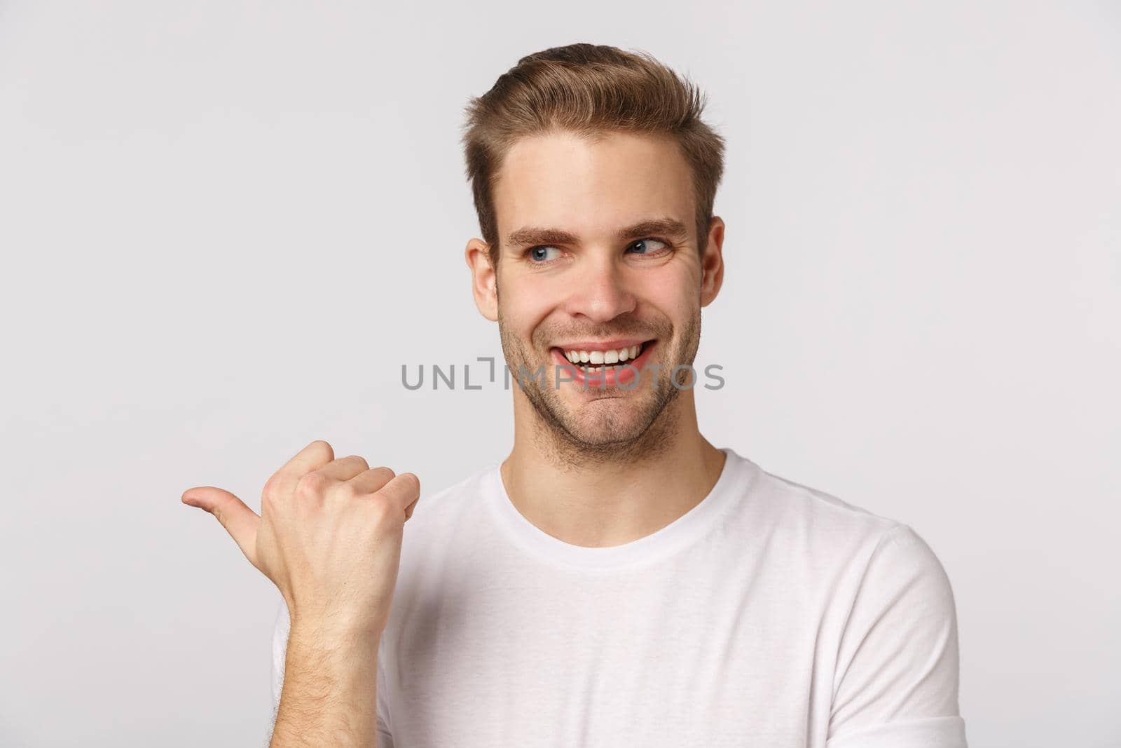 Intrigued and sly good-looking blond bearded guy with bristle in white t-shirt, peeking and pointing left with excited, thrilled smile, standing enthusiastic, check out something desirable.