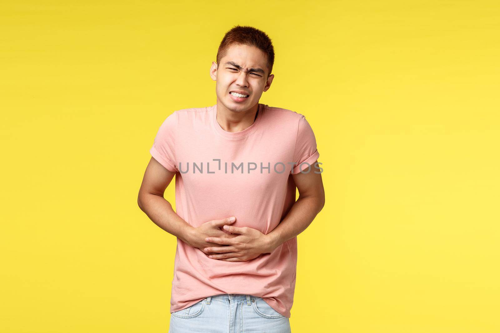 Health, people and food concept. Asian guy in pink t-shirt, feel sick, grimacing from pain in stomach, standing over yellow background suffer from poisoning, hold hands on belly and bending.