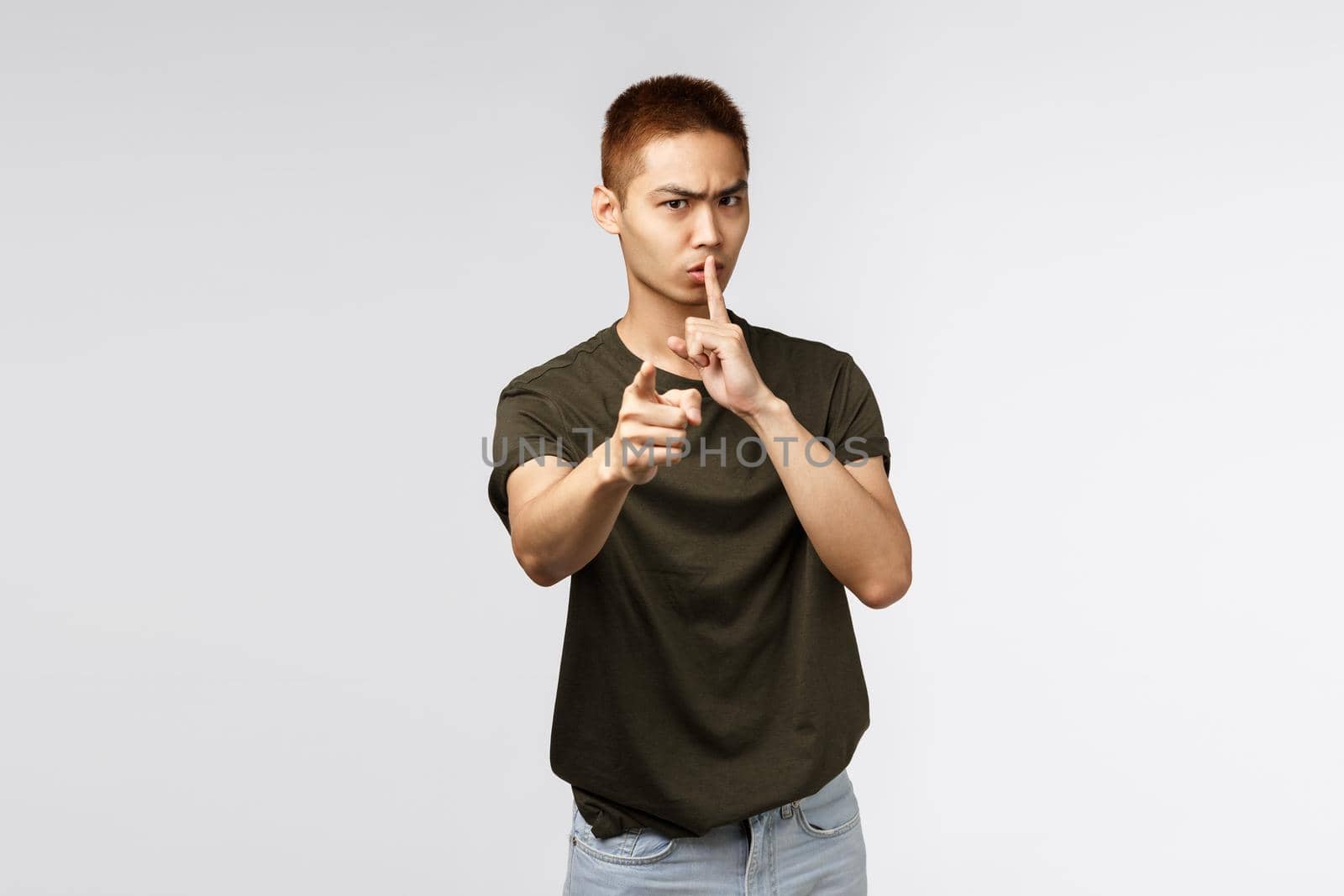 You better keep mouth shut. Portrait of angry serious-looking asian man pointing at camera and shushing scolding you for being noisy and loud, tell person be quiet and dont speek, grey background.