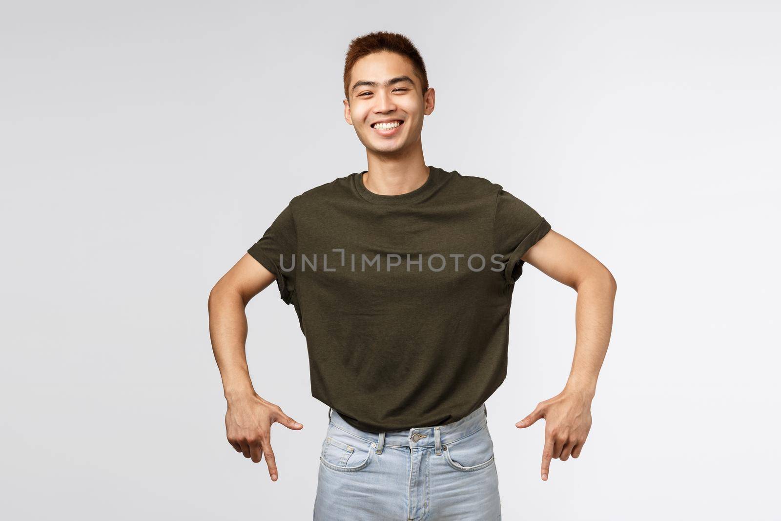 Portrait of cheerful asian man in greey t-shirt, laughing and smiling joyfully, pointing fingers down, invite to click link, subscribe or find out offer details at bottom banner, grey background.