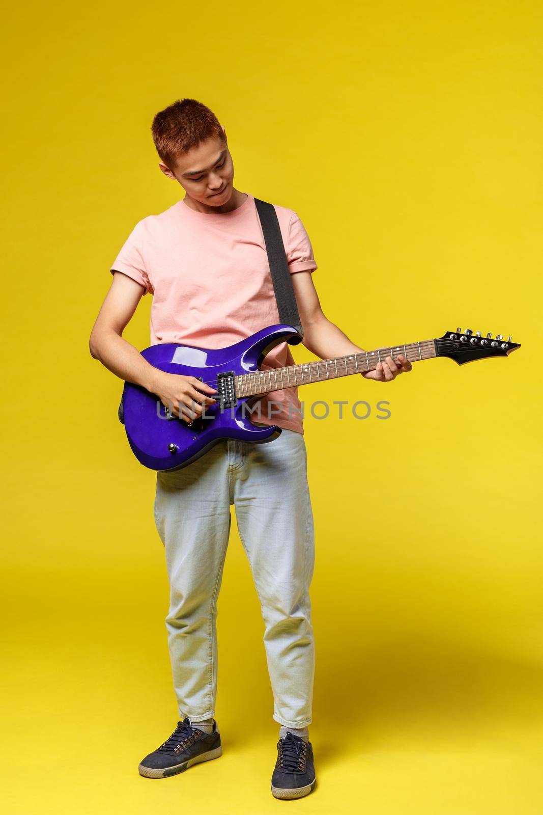 Lifestyle, leisure and youth concept. Vertical shot of creative good-looking asian man playing electric guitar, tuning in before perform on stage, look at instrument, stand yellow background.