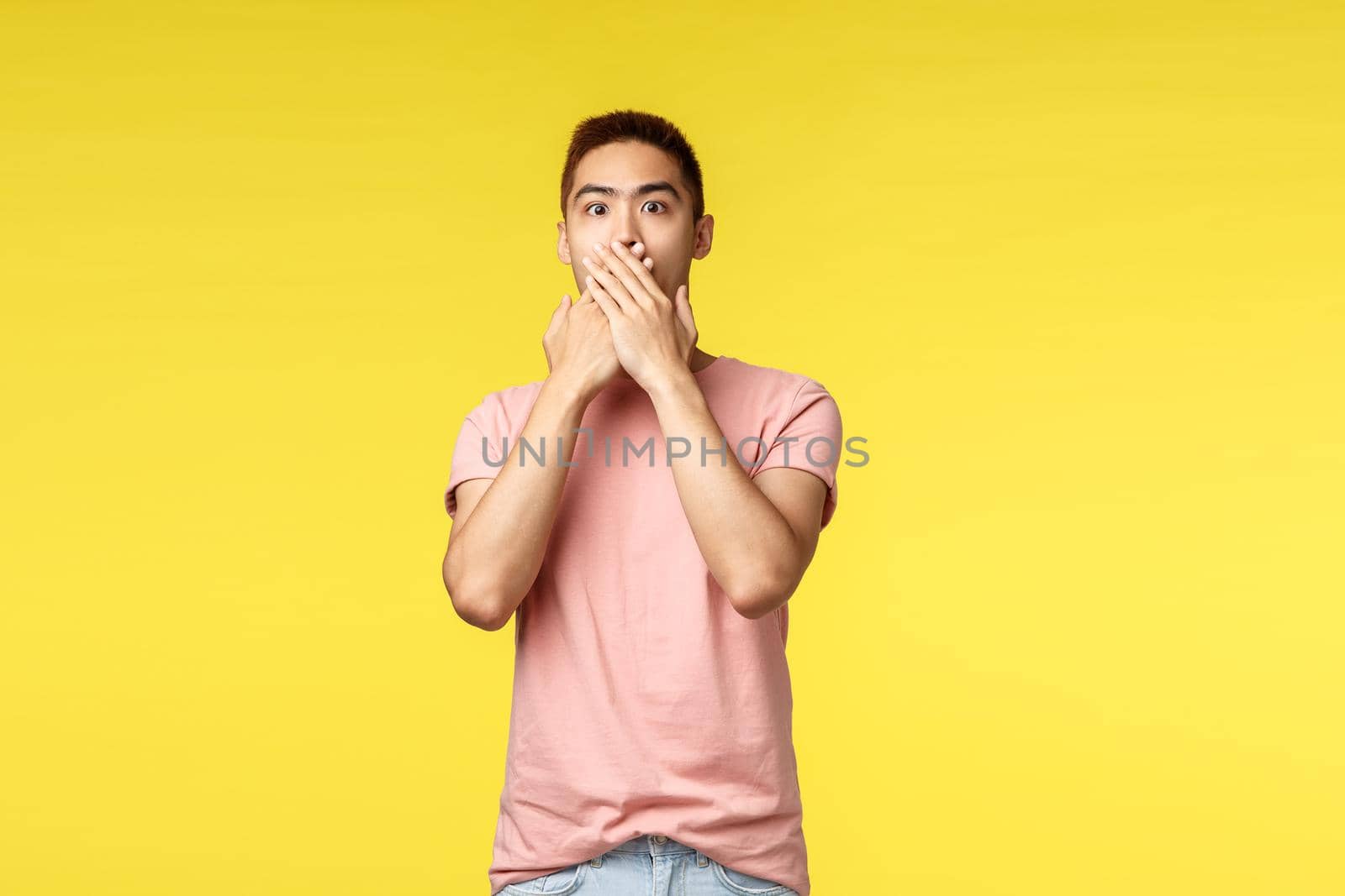 People, different emotions and lifestyle concept. Portrait of shocked asian hipster guy in pink t-shirt, gasping amazed, cover mouth and express amazement with eyes, standing yellow background.